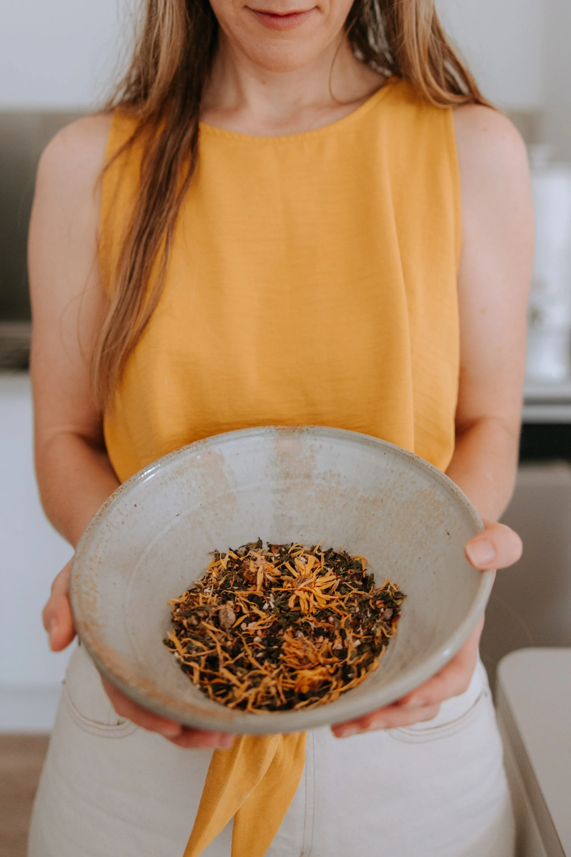 Person in an orange sleeveless top holding a bowl of dried herbal tea or tea leaves.
