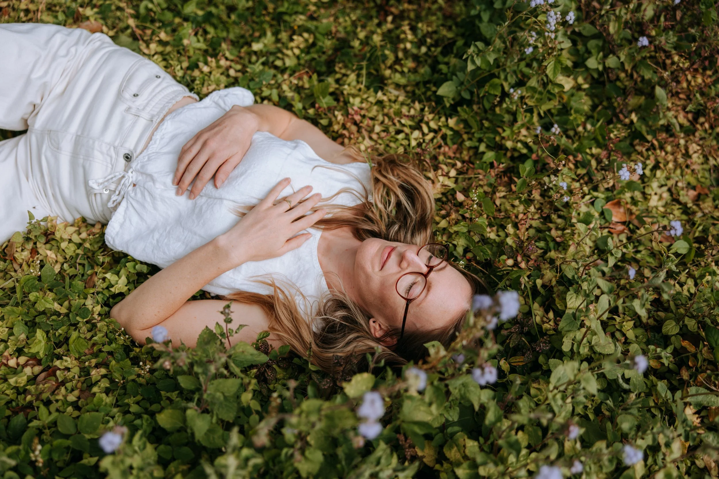 Woman with glasses lying on green foliage with small blue flowers, eyes closed and arms crossed over her chest.