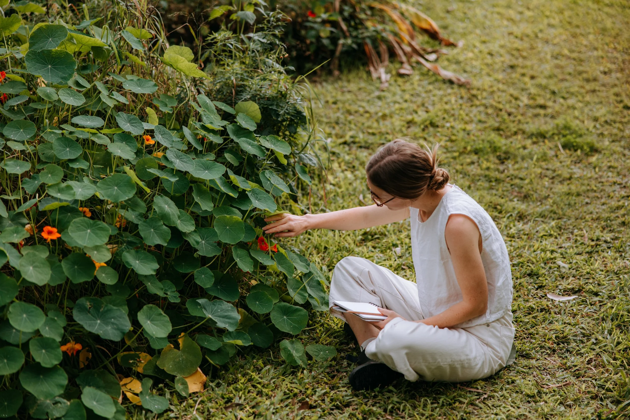 A woman sitting on the grass near a bush of green leaves and orange flowers, wearing a white sleeveless top and beige pants, holding a notebook while examining the plants.