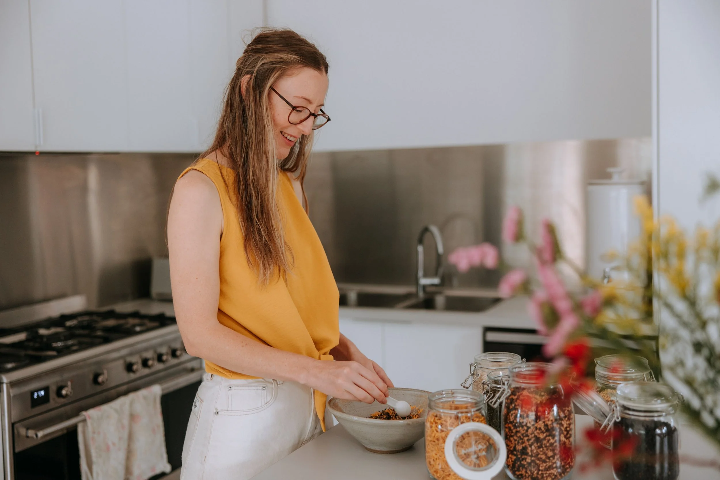 A woman with long hair, glasses, wearing a yellow top, smiling as she prepares food in a modern kitchen with jars of spices on the counter.