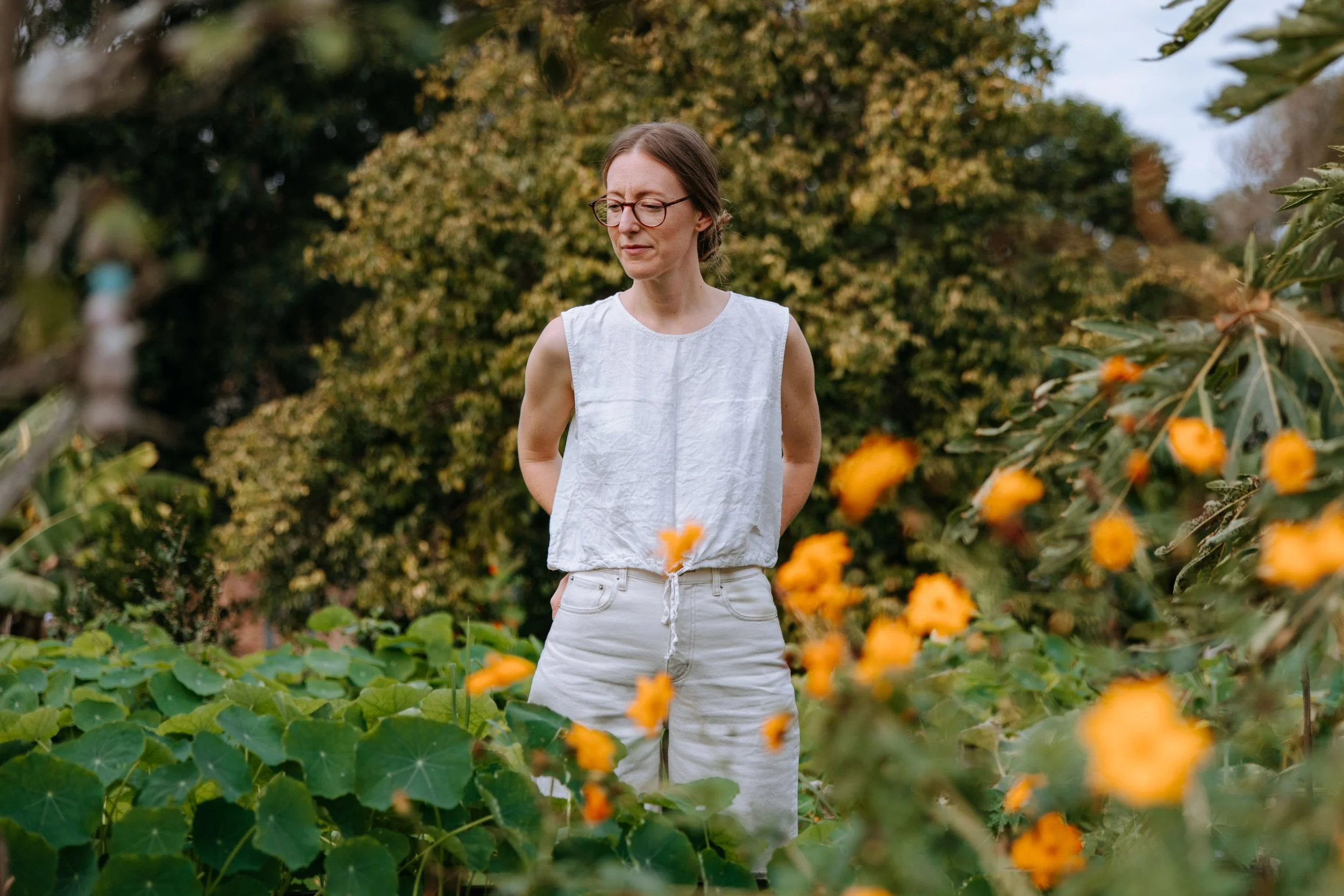 A woman with glasses and light-colored clothing standing in a garden surrounded by orange flowers and green foliage.