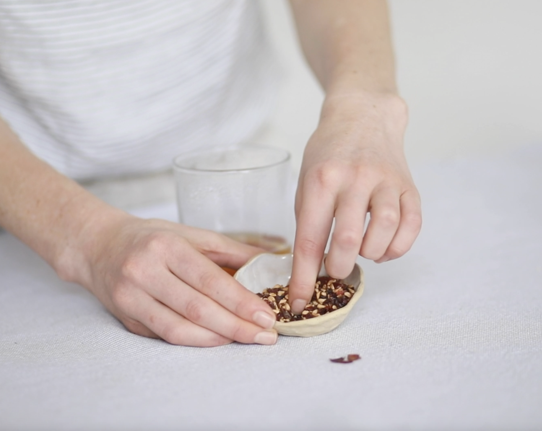 Person's hands scooping crushed red pepper flakes from a small bowl onto a table with a glass of iced tea in the background.