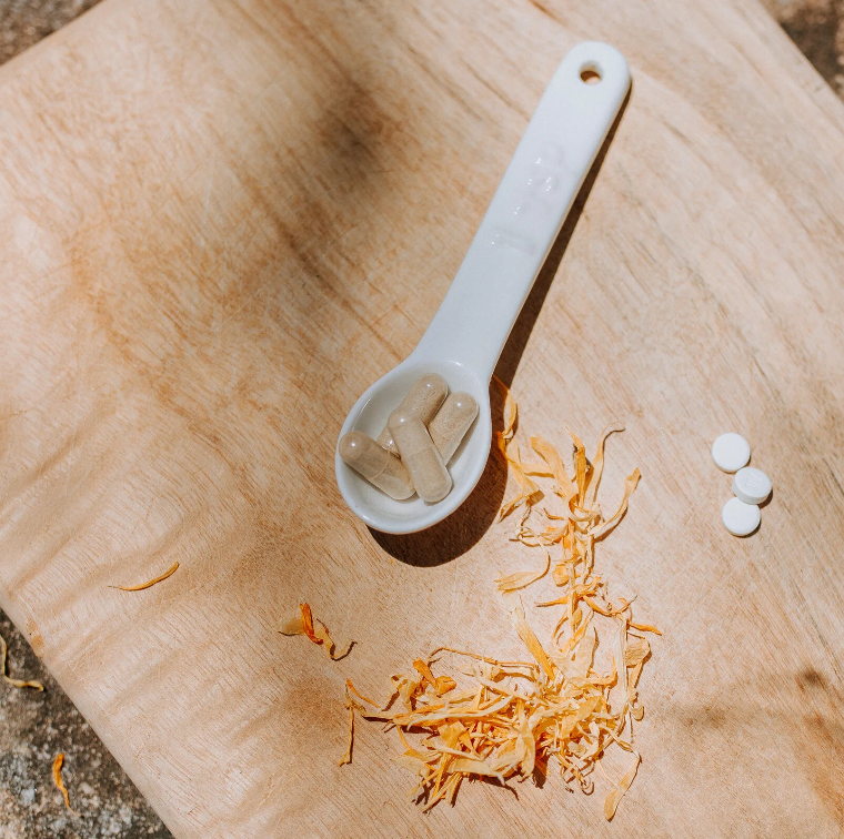 A white ceramic spoon with beige capsules inside, next to scattered shredded paper on a wooden surface, with a few white tablets nearby.