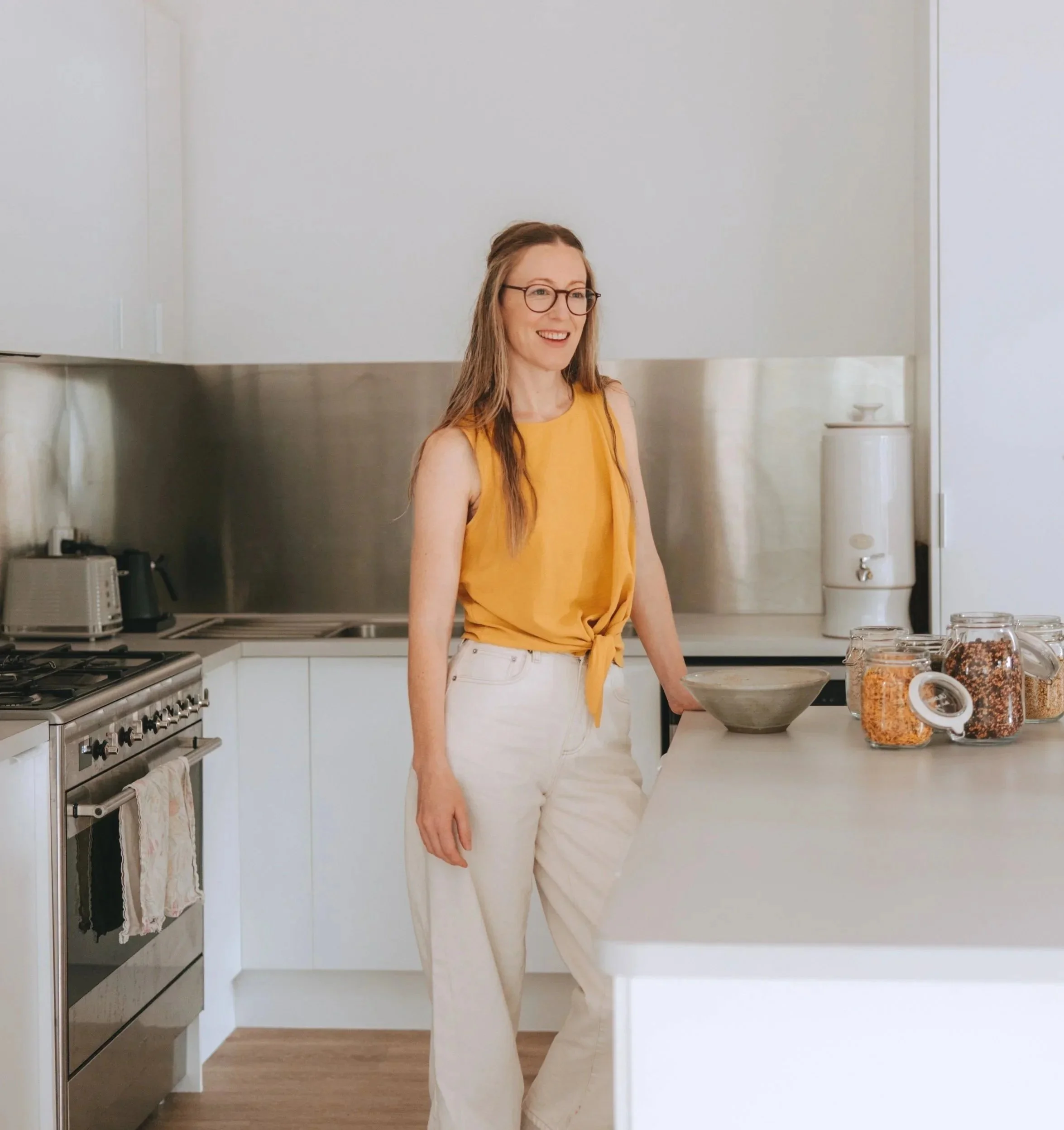 A woman with long hair and glasses standing in a modern kitchen, smiling, wearing a yellow sleeveless top and white pants, next to jars of granola on a white counter.
