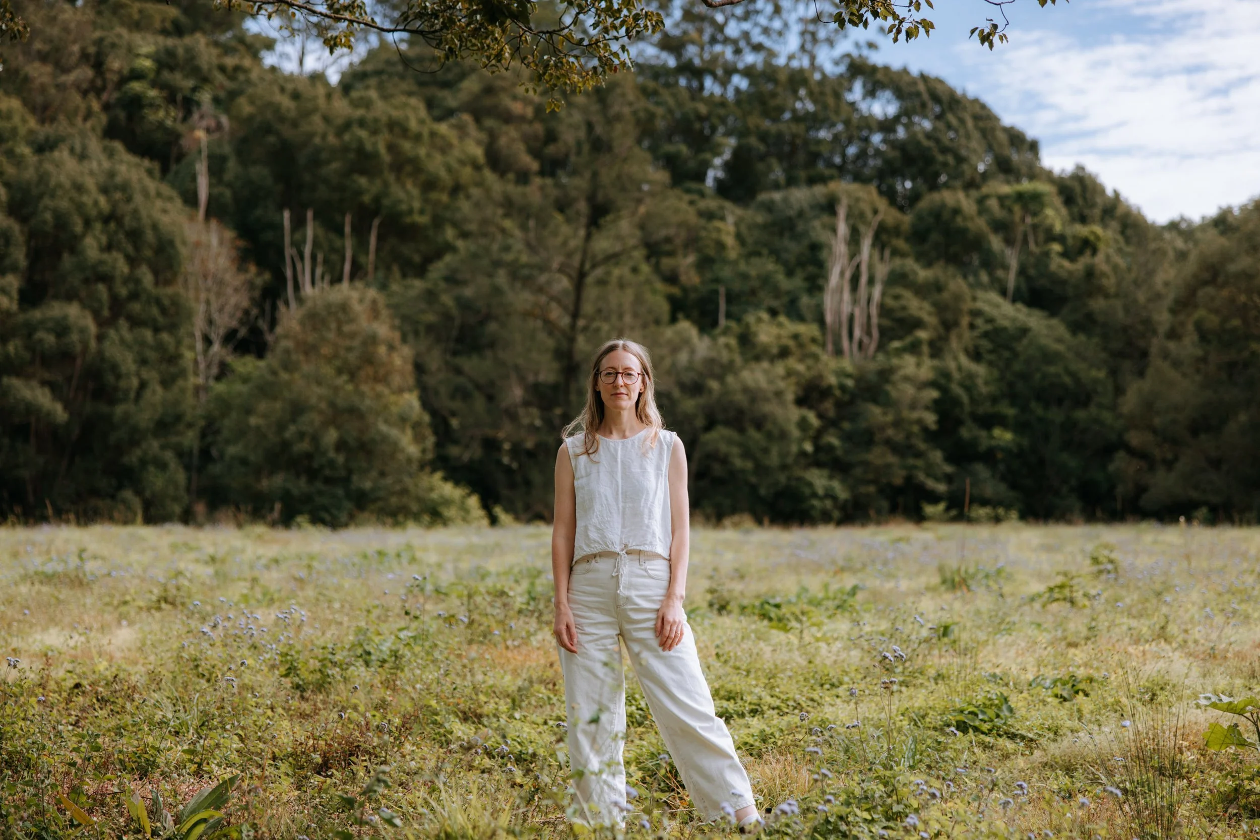 A woman standing in a grassy field with a backdrop of trees and hills under a partly cloudy sky.