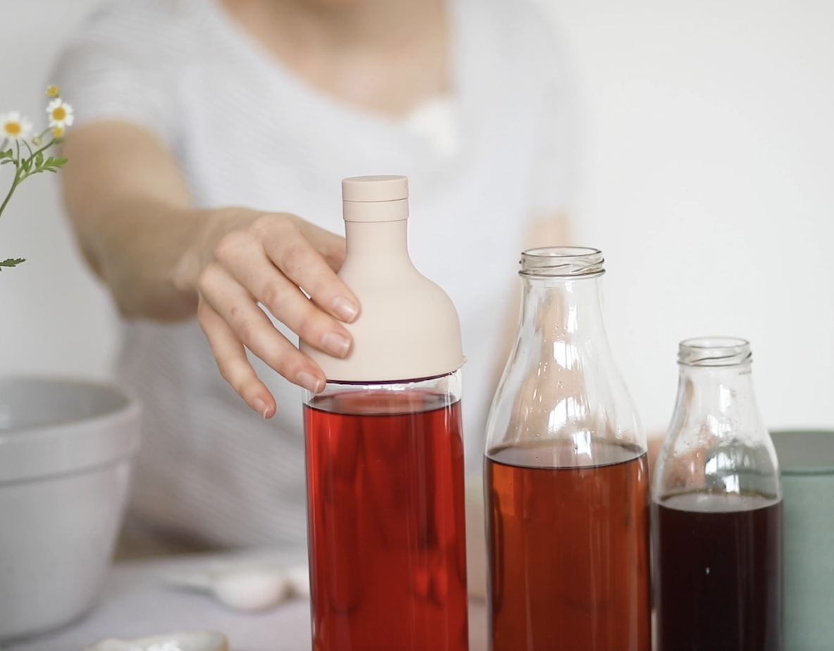 A person pouring red liquid from a bottle with a beige cap, surrounded by other glass bottles of amber liquid, on a table with a gray bowl and a plant with small white and yellow flowers.