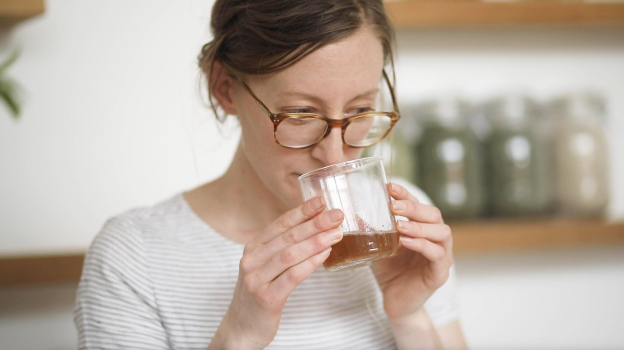 A woman with glasses drinking a glass of dark-colored beverage in a bright kitchen.