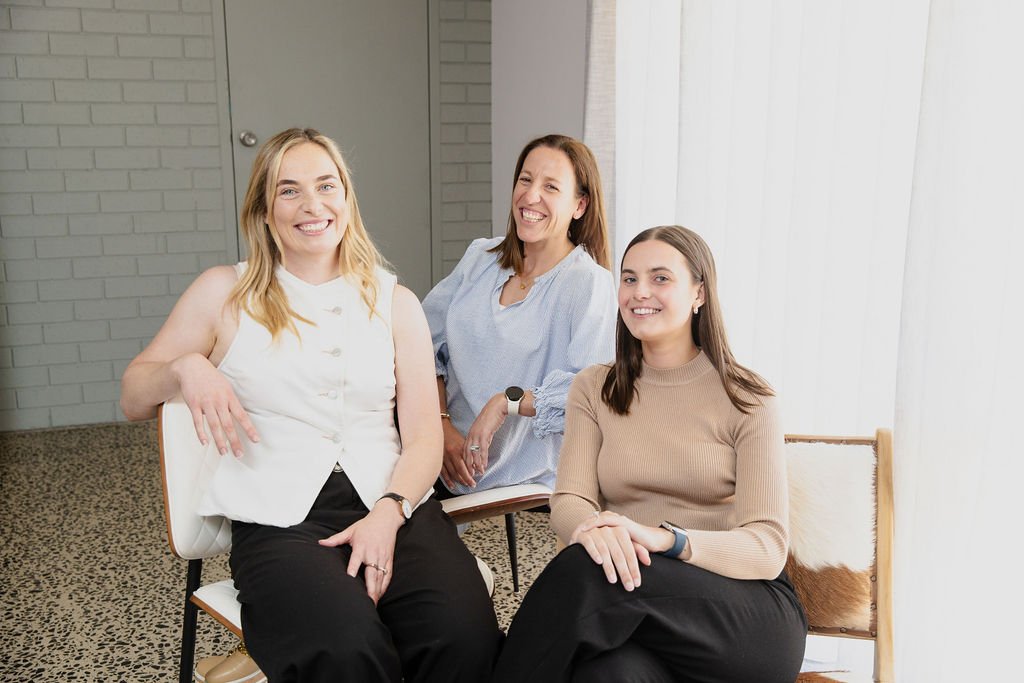 Three women seated in a well-lit room, dressed in professional attire, smiling at the camera.