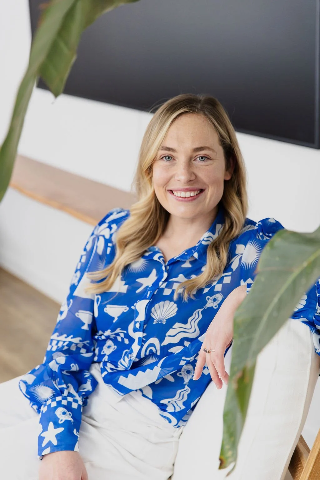 Imogen - a miling woman sitting, wearing a blue patterned blouse, framed by green leaves.