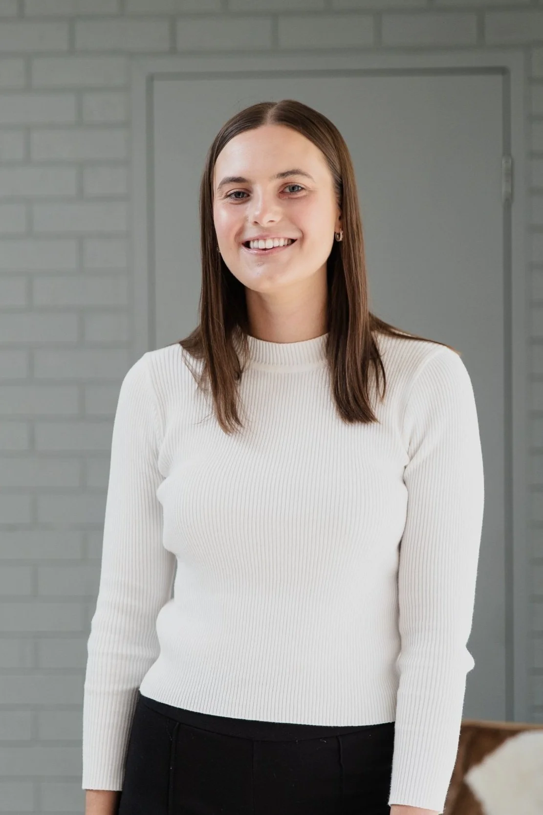 Grace - a smiling woman in a white sweater standing against a gray brick wall.