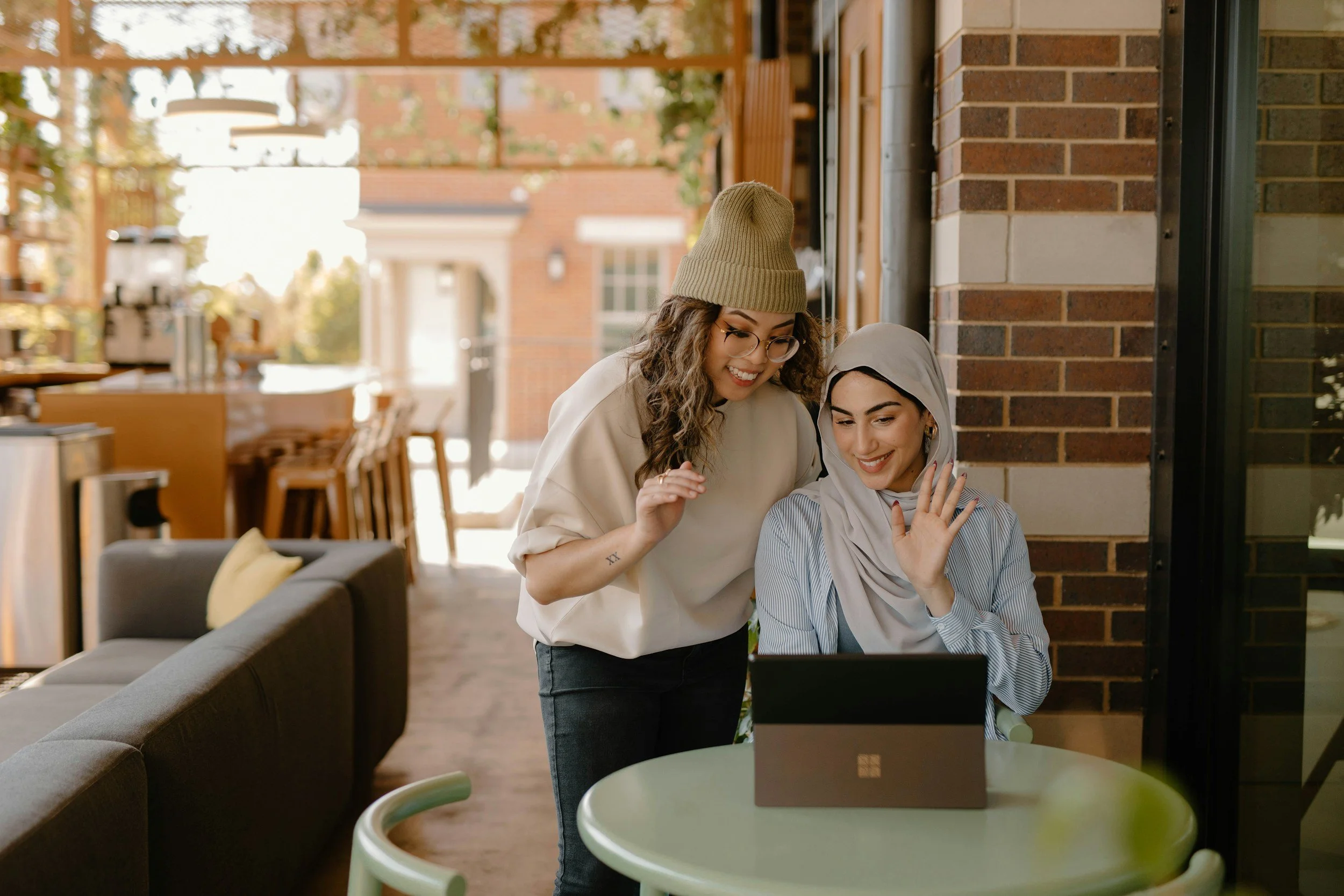 Two women, one wearing a hijab and the other wearing a beanie, look at a laptop together in a cozy indoor café or restaurant, smiling and waving.
