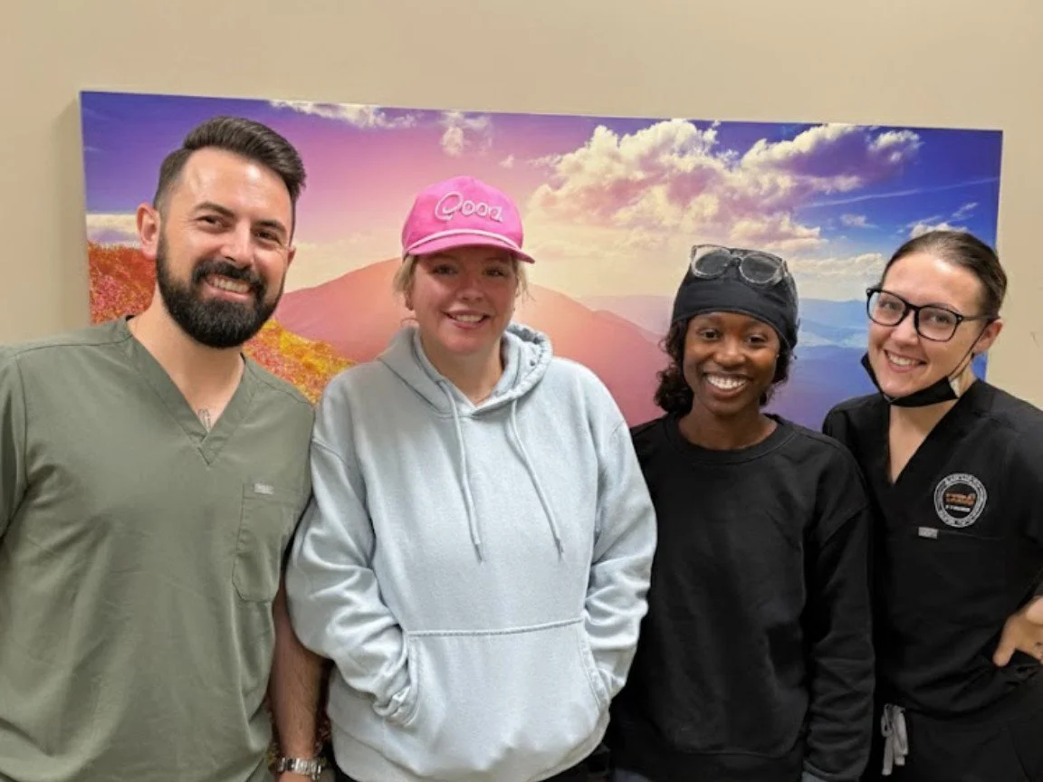 Four smiling people standing in front of a landscape picture with mountains and a colorful sky.