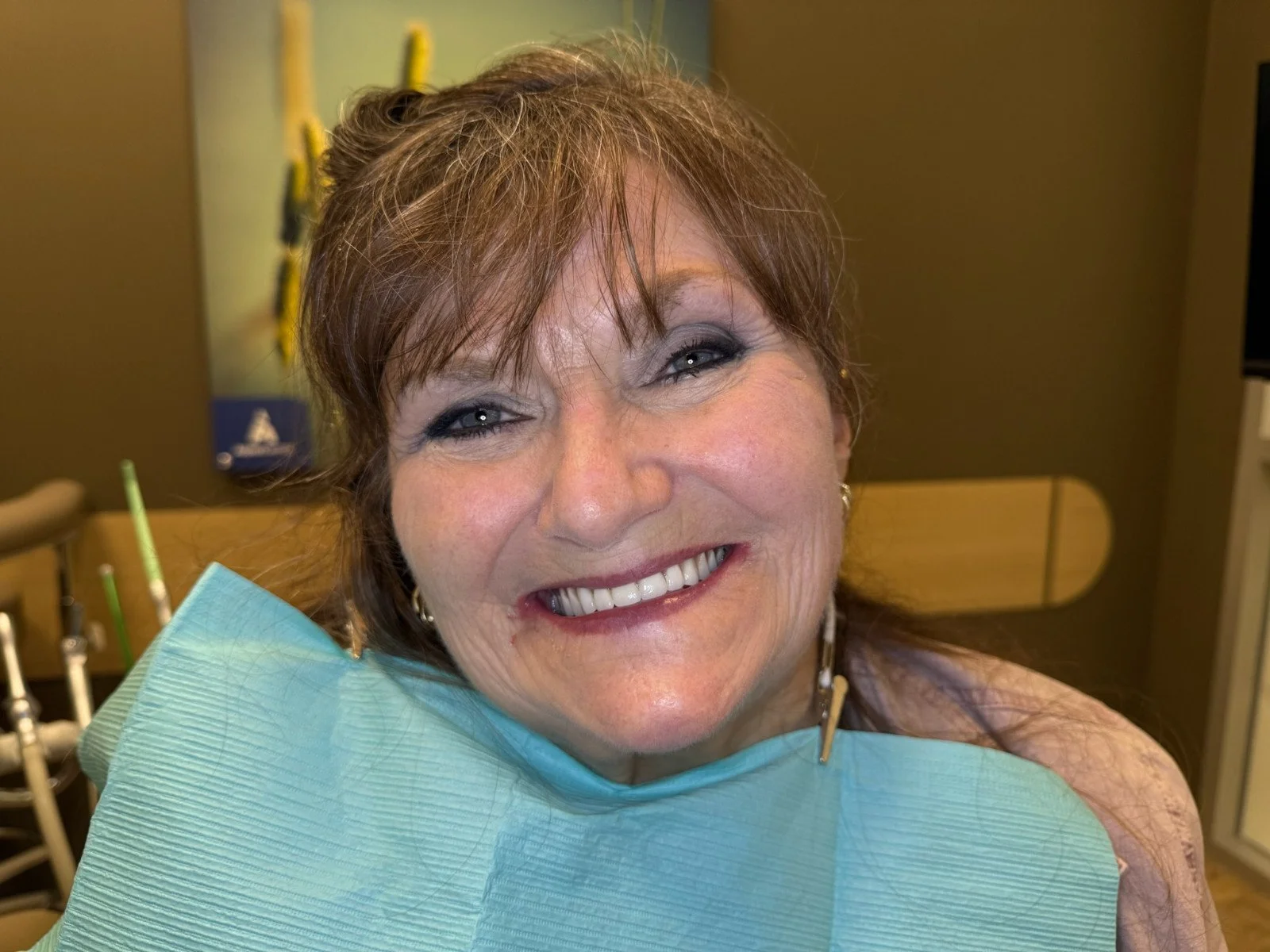 Close-up of a smiling middle-aged woman with short reddish-brown hair, makeup, earrings, and a bib, sitting in a restaurant or cafe.