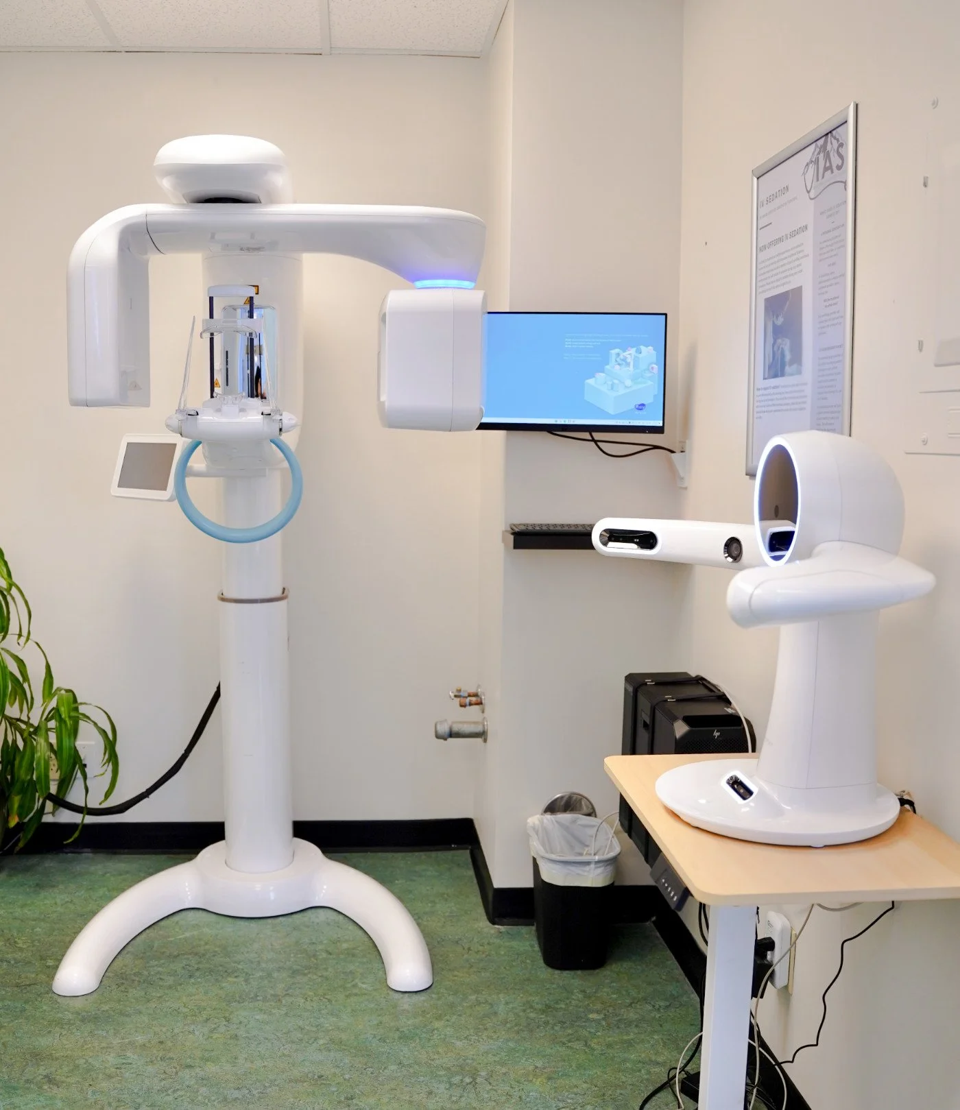 Dental imaging equipment including a panoramic X-ray machine, a wall-mounted monitor, and a secondary imaging device on a small table in a room with green carpet and a plant.