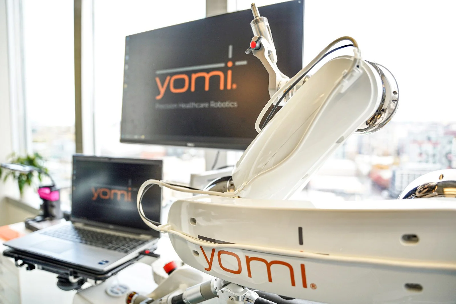 Robotic medical device with the logo 'Yomi', placed in a medical setting with screens and a laptop in the background.