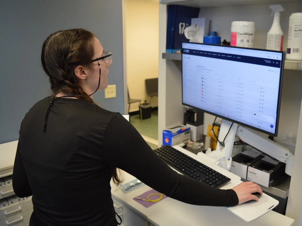 A woman with glasses and braided hair working on a computer in a medical or office setting, with shelves containing medical supplies and bottles behind her.