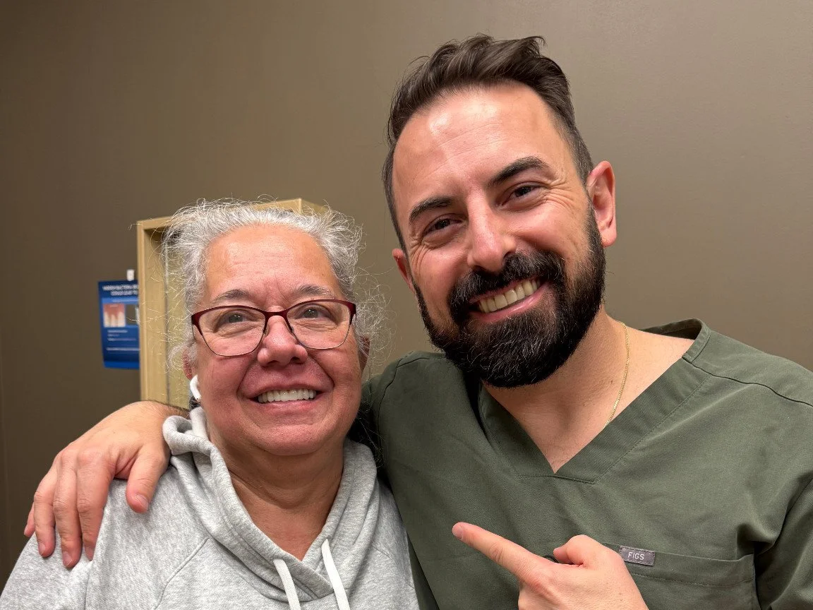 A woman with gray hair, glasses, and a gray hoodie smiling and posing with a man with dark hair, beard, scrubs, and a gold necklace, smiling and pointing at her in an indoor setting.
