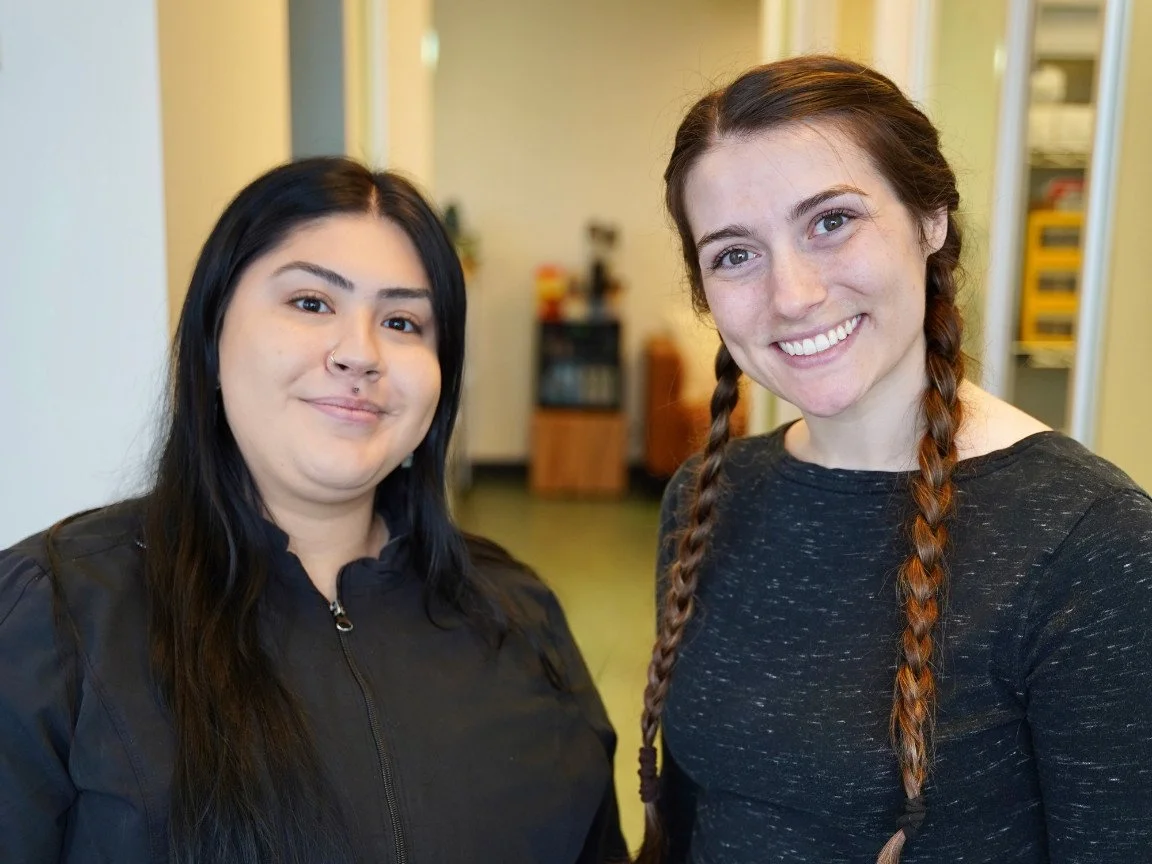 Two women smiling indoors, one with dark hair and a nose piercing, the other with red hair in braids, both wearing dark tops.