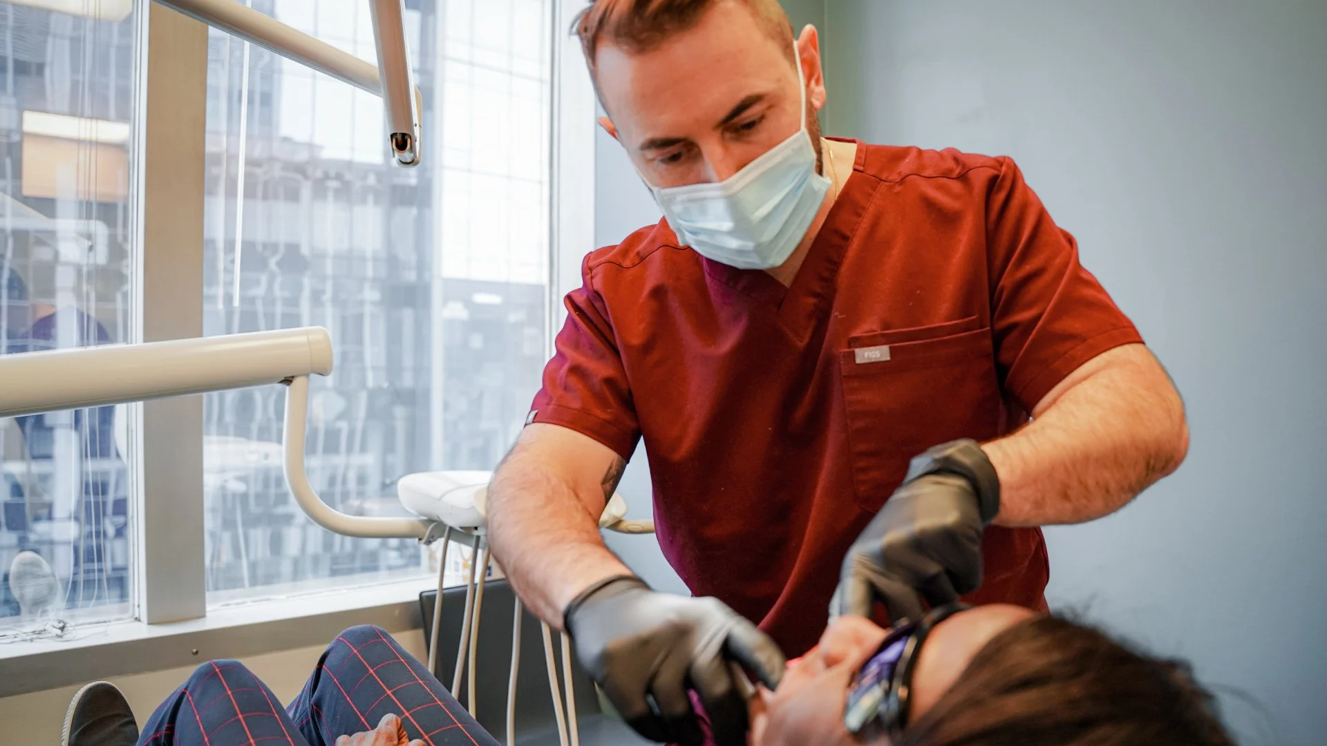 A healthcare professional wearing a mask, gloves, and red scrubs attending to a patient lying on a hospital bed.