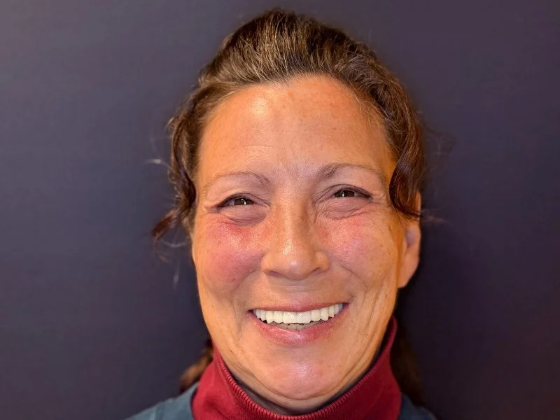Close-up of a smiling woman with light skin, brown hair, and wearing a red and black shirt, against a dark background.