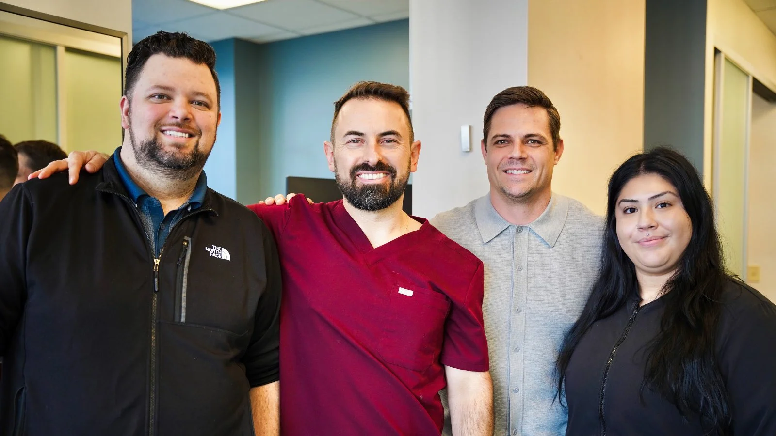 Four people standing together indoors, smiling at the camera, with one person wearing a North Face jacket, a person in a red medical uniform, and others in casual clothing.