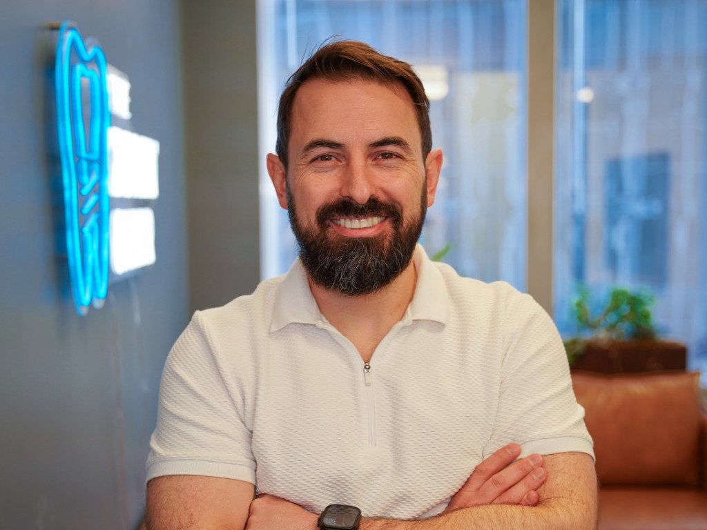 A smiling man with a beard and mustache wearing a white polo shirt, standing indoors with arms crossed, in front of a glass window and blue neon sign.