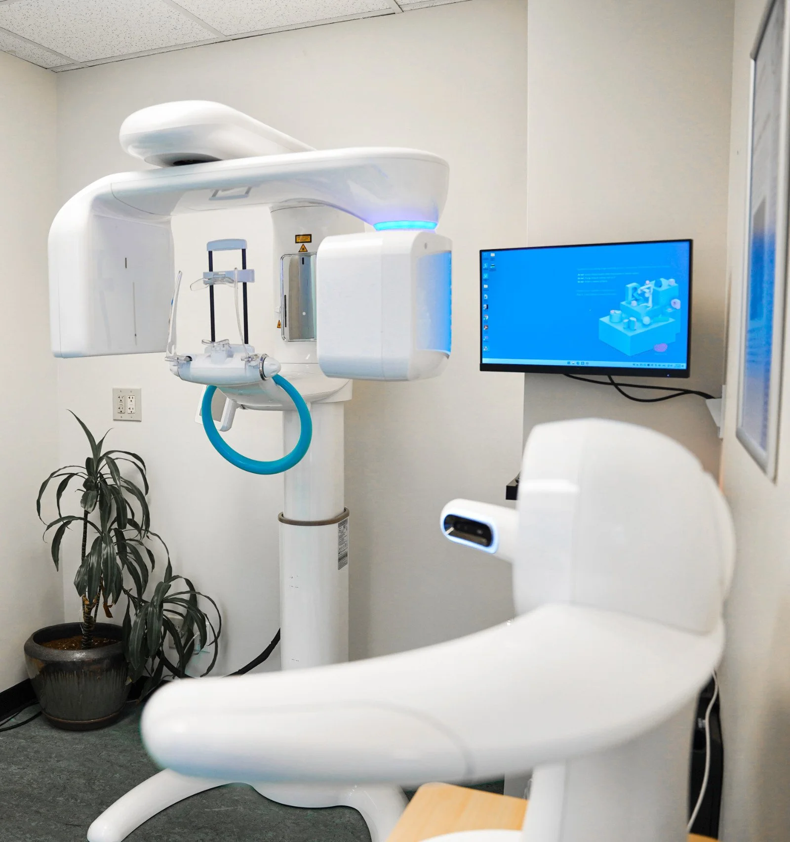 Dental implant imaging room with a large imaging machine, a potted plant, and a wall-mounted monitor displaying a blue screen.