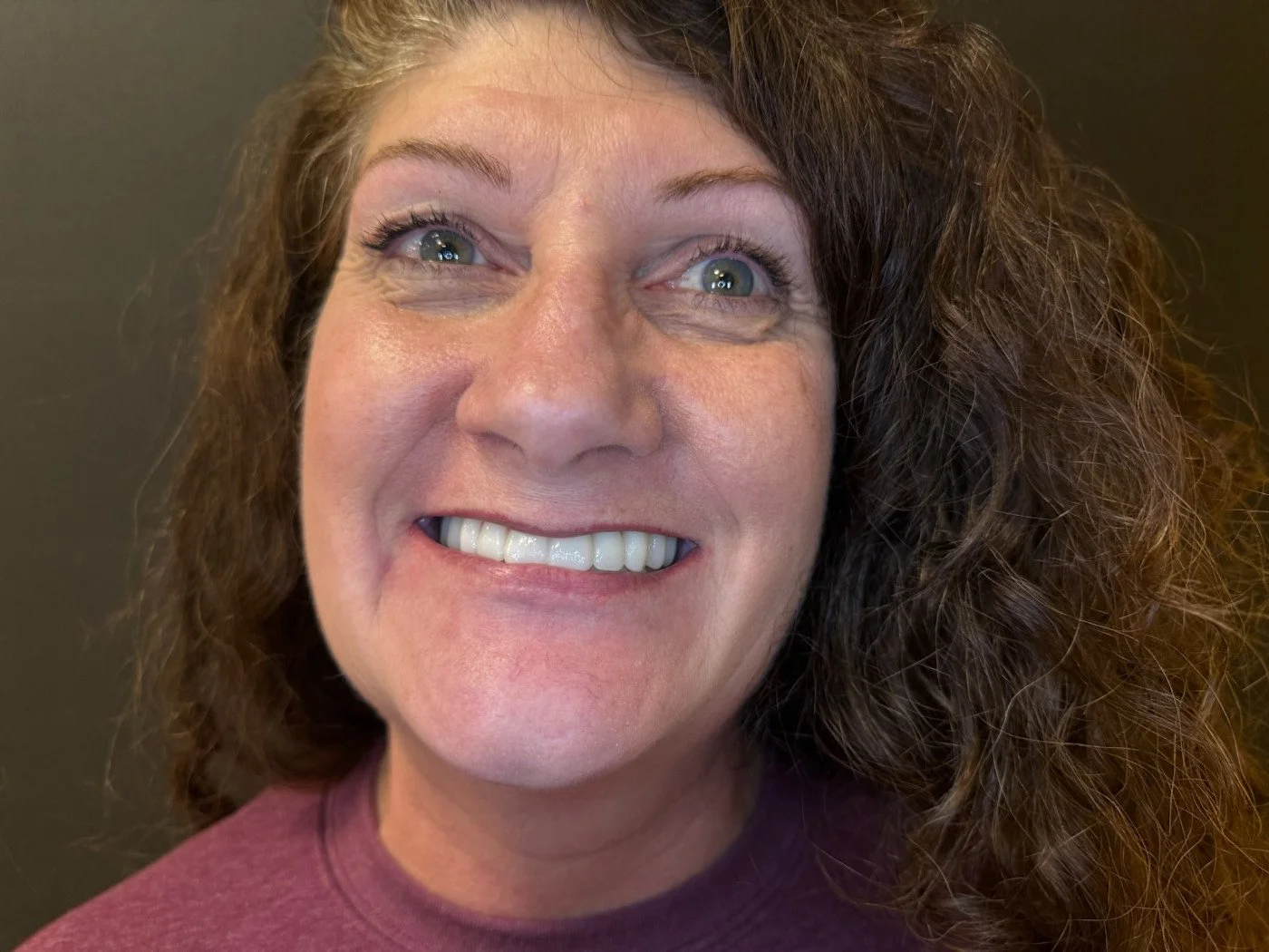 Close-up portrait of a smiling woman with curly brown hair and blue eyes wearing a maroon top, looking up slightly.