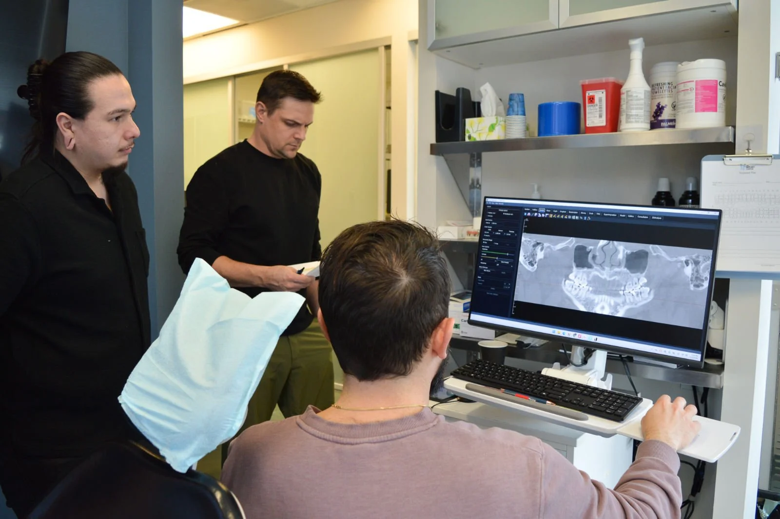 Group of three people, two standing and one sitting, looking at a computer screen displaying a dental X-ray, performing a dental implant consultation.