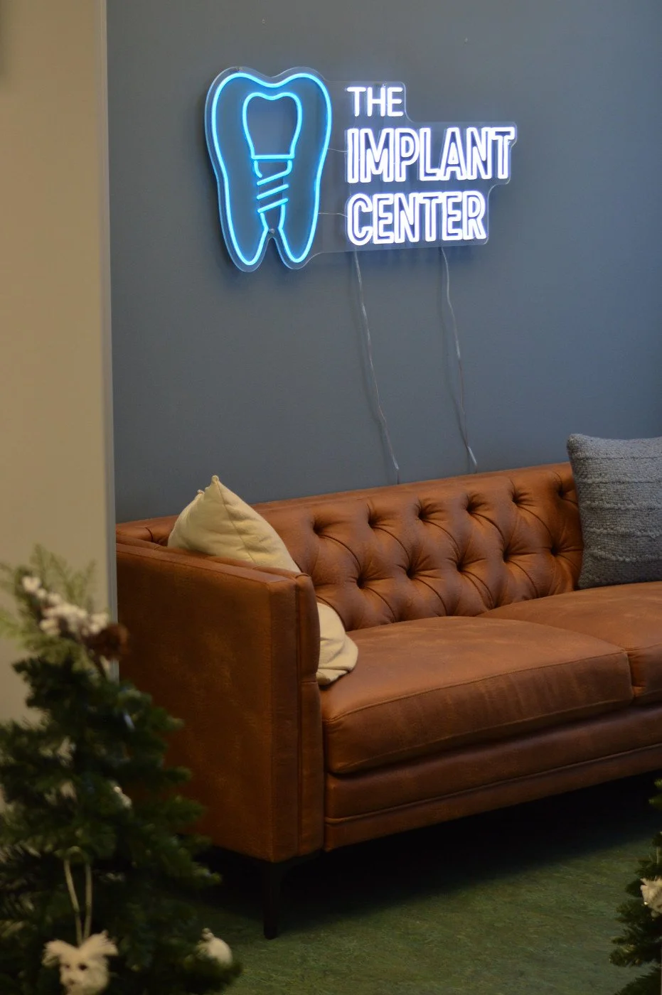 Neon sign reading 'The Implant Center' with a tooth and implant illustration on a gray wall above a brown leather couch in a waiting room.
