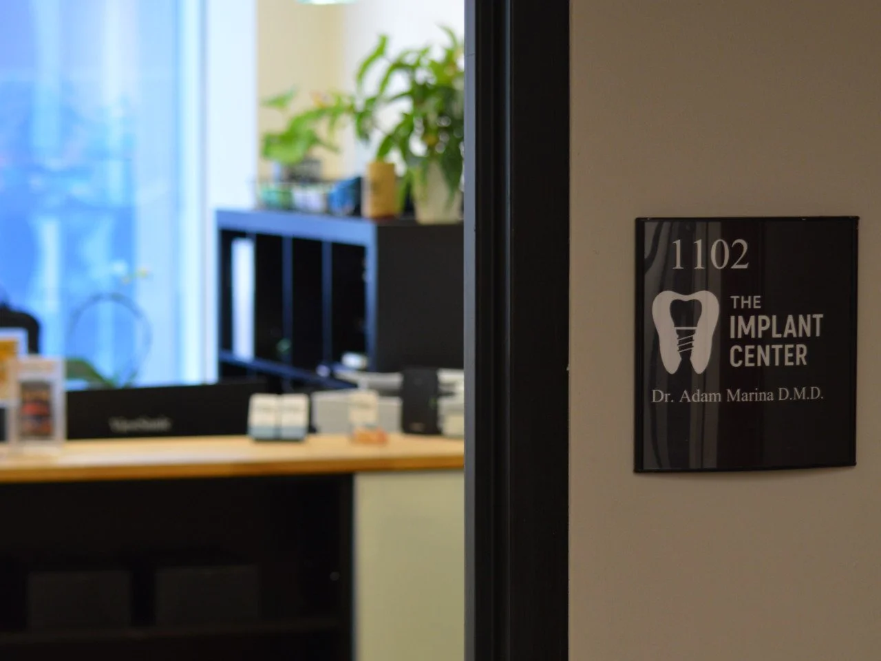 Office entrance sign for the Implant Center with the name Dr. Adam Marina D.M.D., near a room numbered 1102, with an illustration of a tooth with a dental implant.