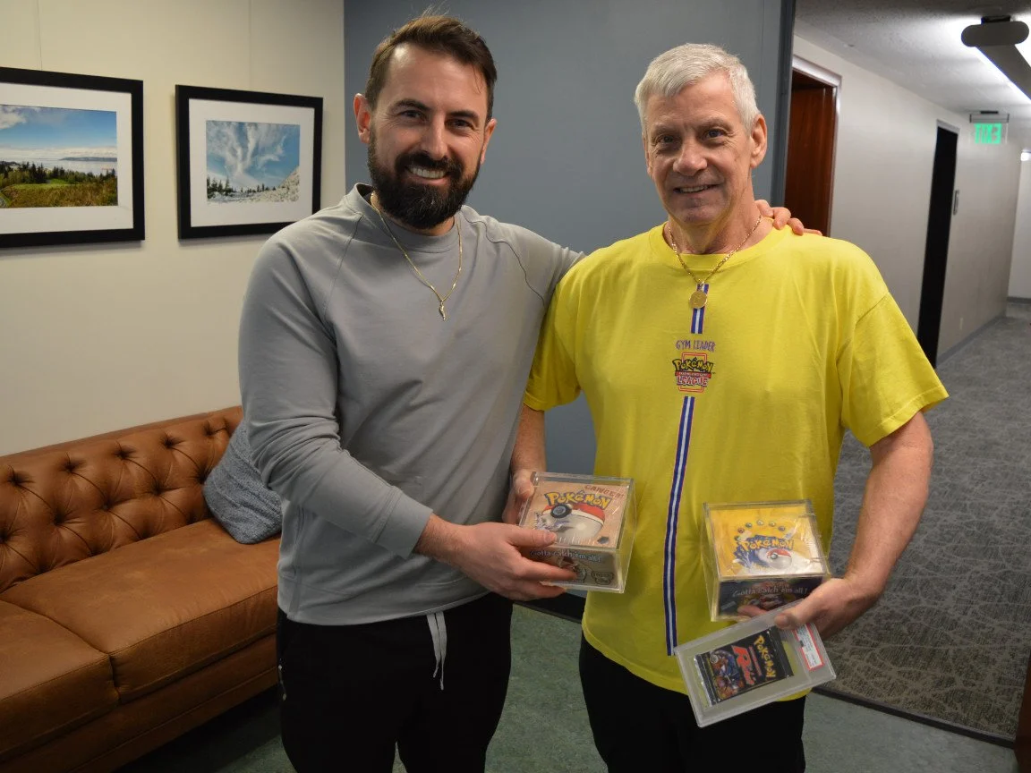 Two men standing together indoors, smiling, each holding Pokémon game boxes in their hands. The man on the left has a beard, is wearing a gray long-sleeve shirt, and has a necklace. The man on the right has gray hair, is wearing a yellow T-shirt with