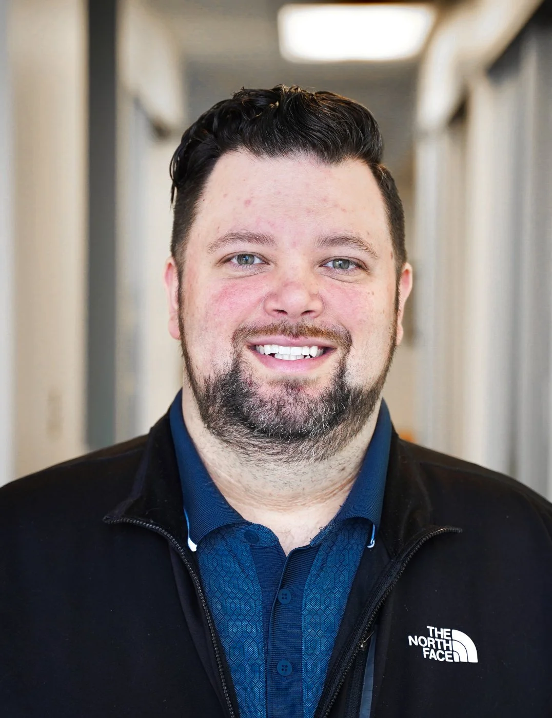Portrait of a smiling man with short dark hair, beard, and blue eyes, wearing a navy blue collared shirt and a black The North Face jacket, standing in a hallway.