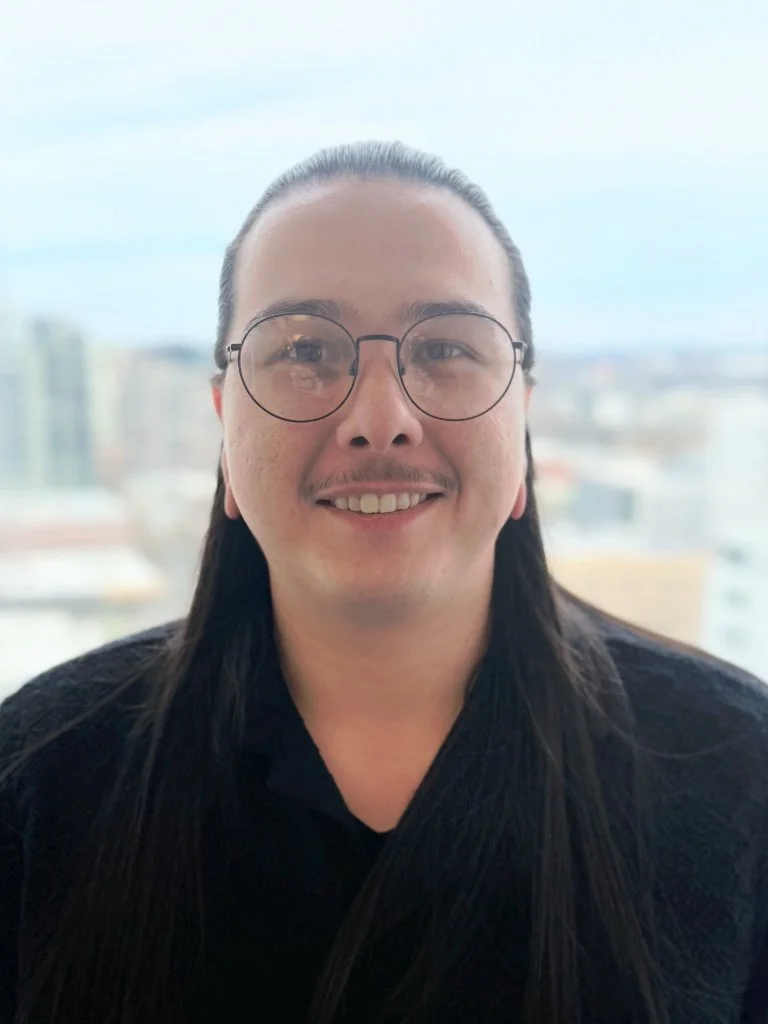 A smiling man with long dark hair, glasses, and a mustache, wearing a black shirt, standing in front of a cityscape background.