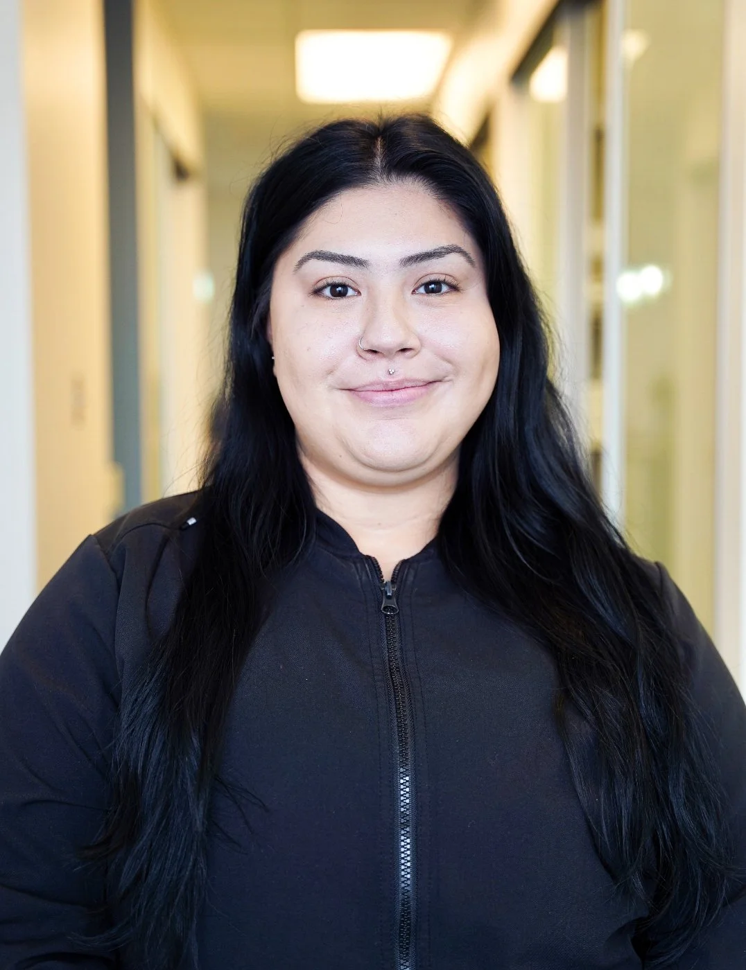 A woman with long black hair smiling in a hallway.