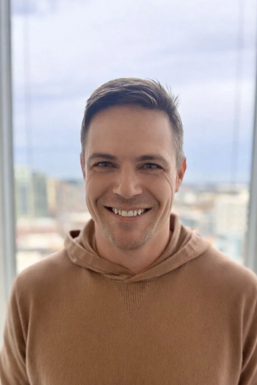 Close-up of a smiling man with short brown hair, wearing a tan hoodie, standing in front of a large window with city buildings and a cloudy sky in the background.