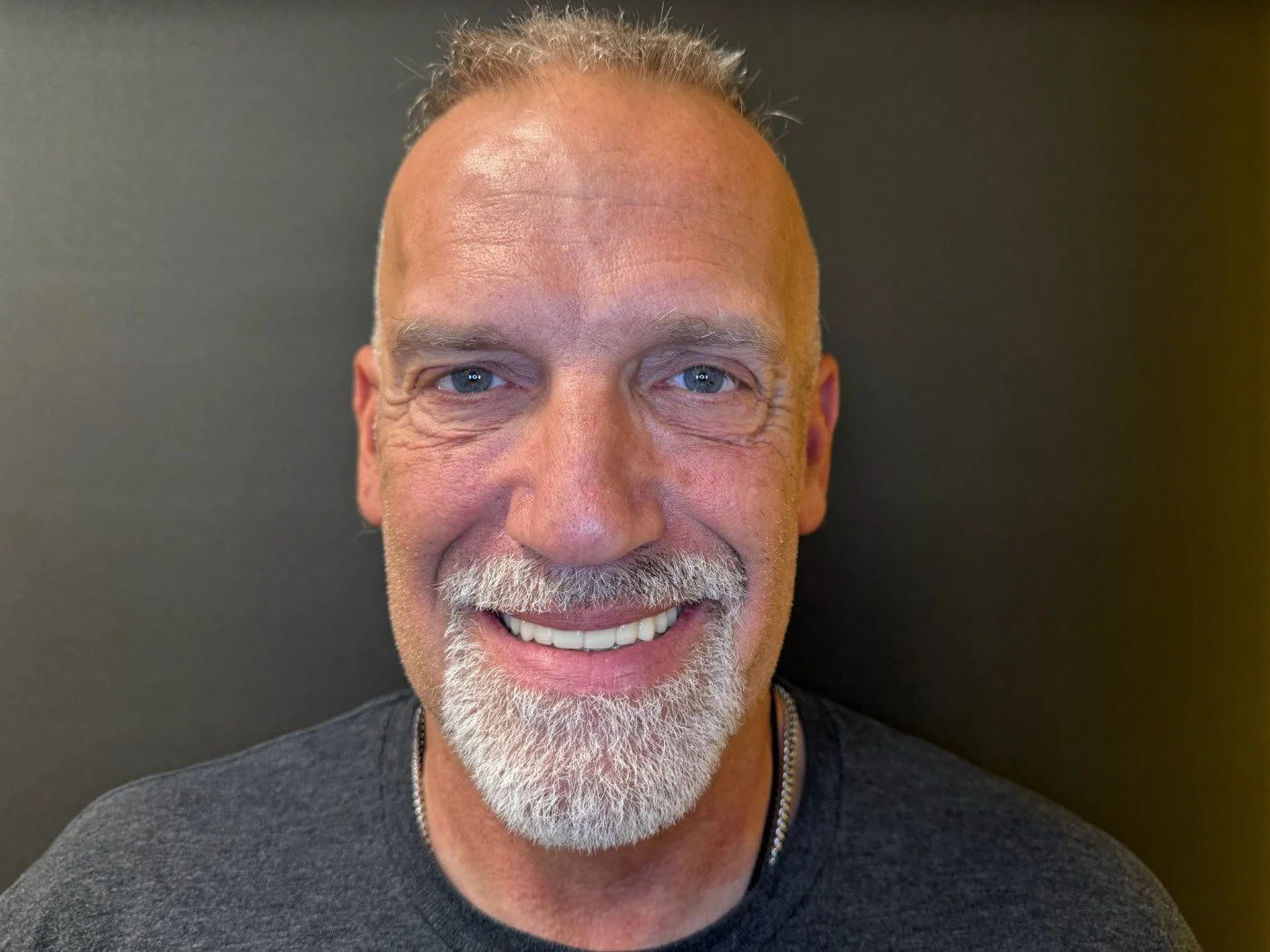 Close-up of a smiling middle-aged man with short gray hair, blue eyes, a gray and white beard, wearing a dark gray shirt, standing against a dark background.