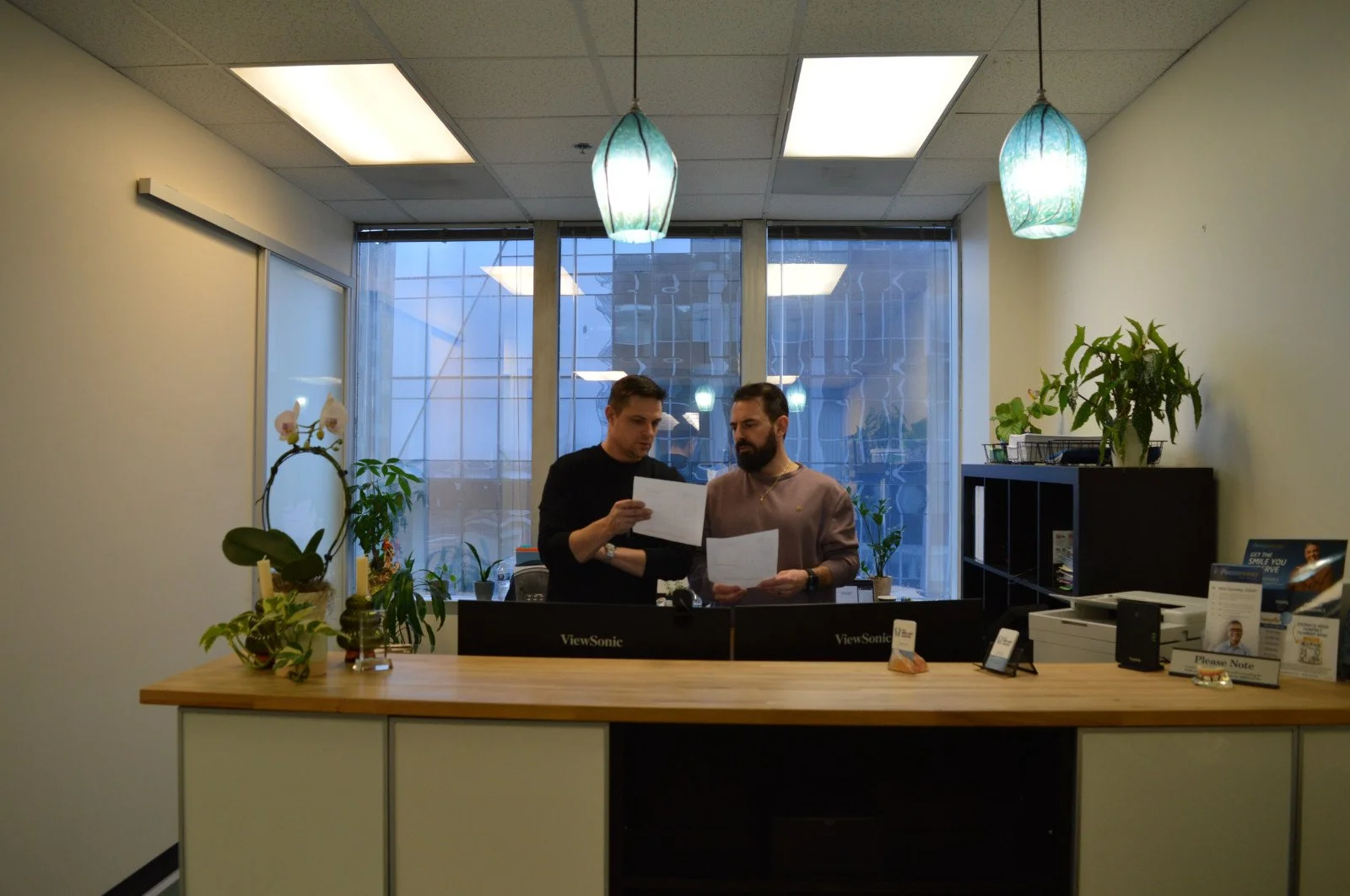 Two men standing behind a reception desk, looking at papers, in an office with plants, a large window, and hanging pendant lights.