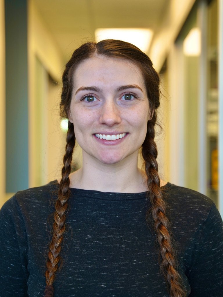A young woman with long braided hair, smiling, standing in a brightly lit hallway.