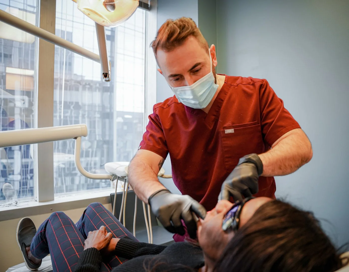 A dental implant specialist wearing a face mask and gloves attending to a patient in a procedure room, with large windows and downtown Portland buildings visible in the background.