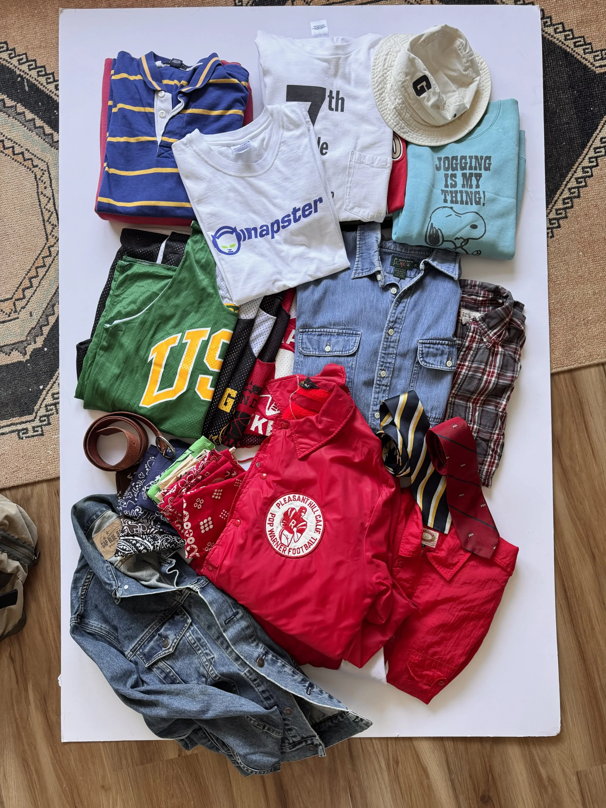 Clothing items including a striped polo shirt, a white T-shirt, a light blue T-shirt with a Snoopy graphic, a white bucket hat, a red jacket, a denim jacket, shorts, bandanas, and a leather belt are spread out on a white surface on a hardwood floor.