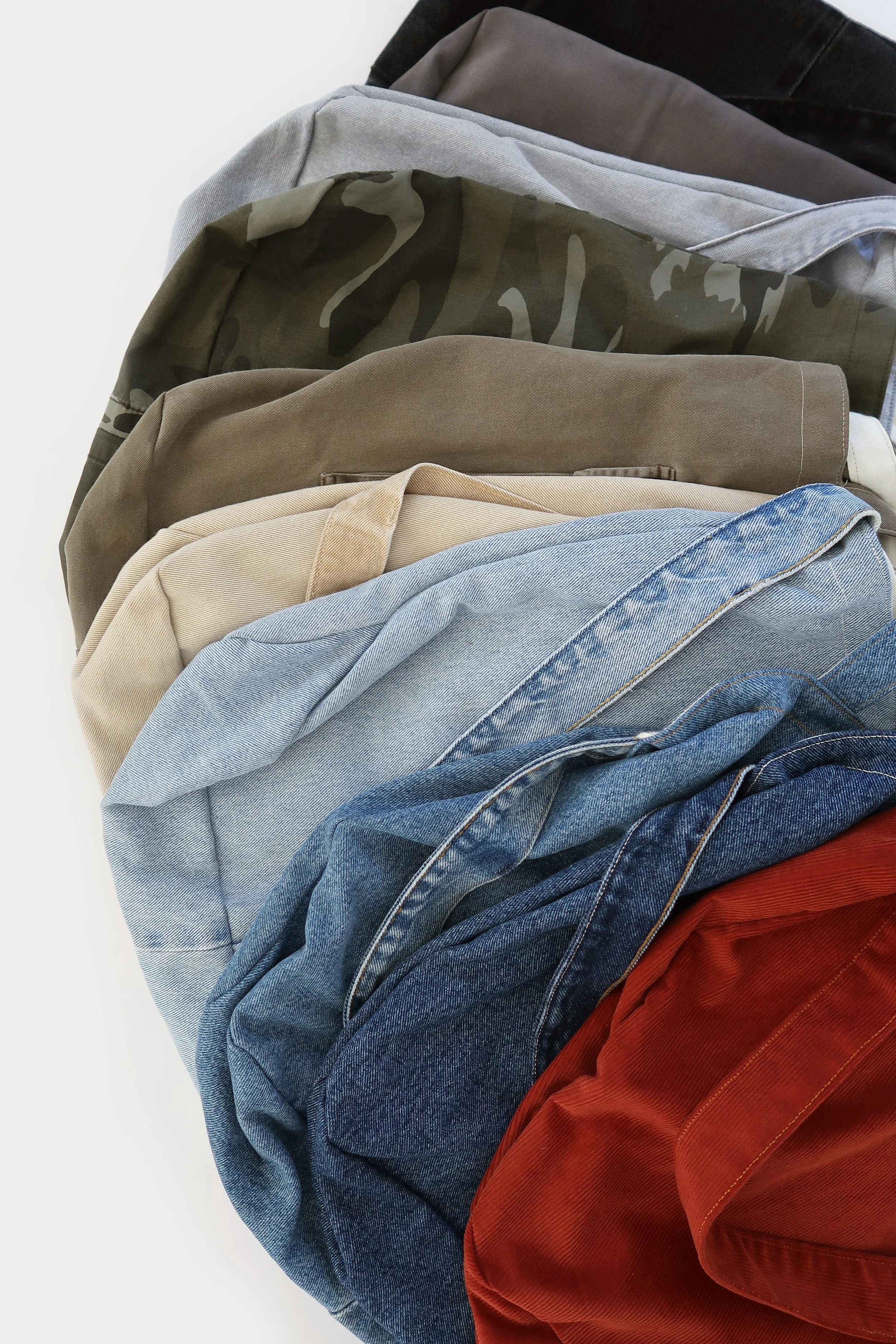 A collection of folded clothing items, including jeans, khakis, camouflage-patterned fabric, and various shirts, arranged overlapping on a white surface.