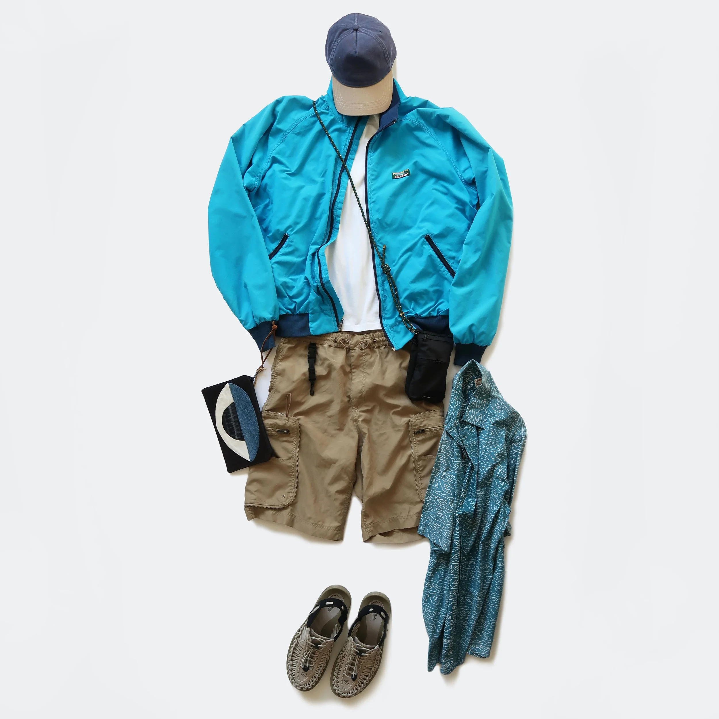 A flat lay display of outdoor and athletic clothing and footwear, including a blue windbreaker, khaki pants, a baseball cap, a pair of hiking shoes, a pair of water shoes, and a blue patterned headscarf, arranged on a white background.