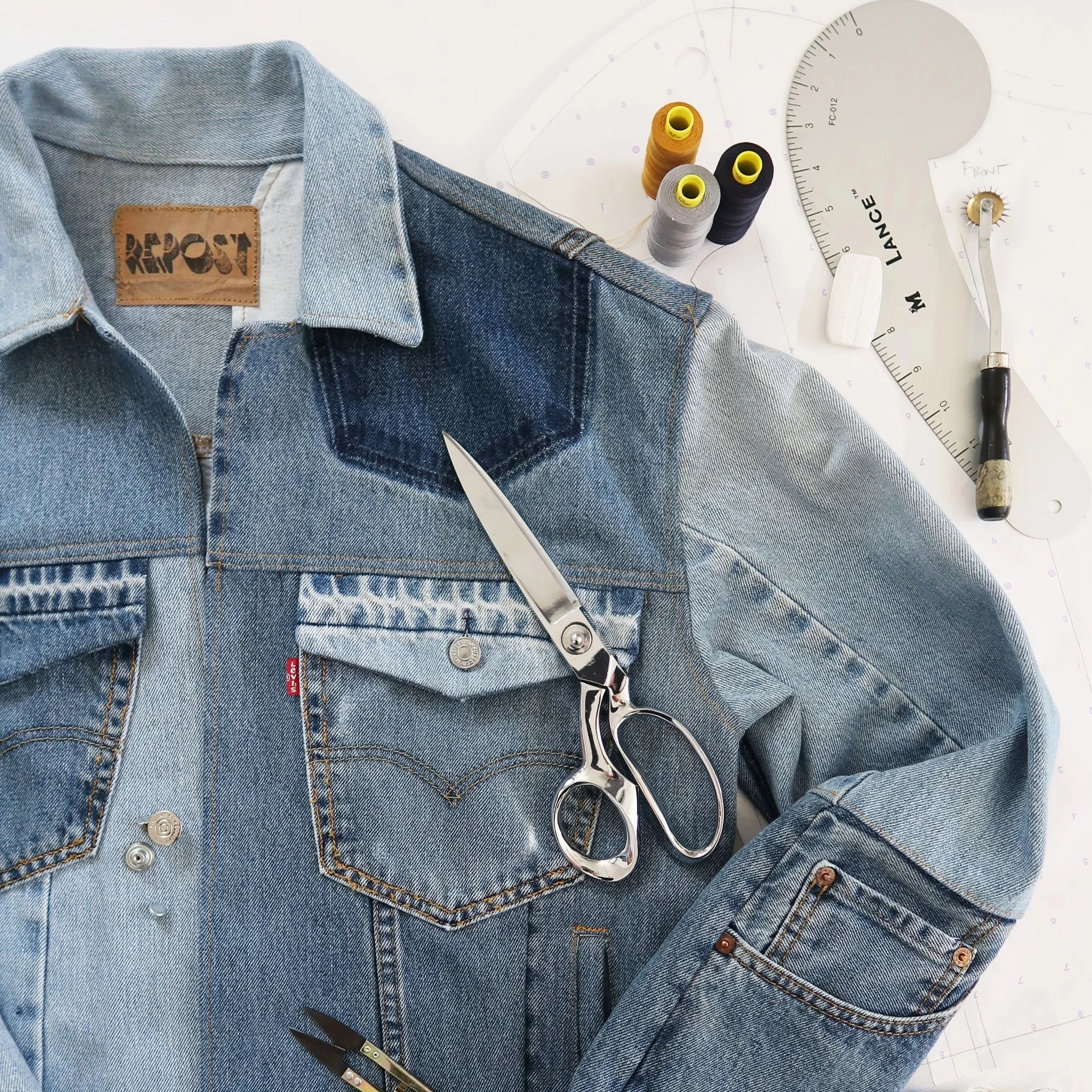 A close-up view of a denim jacket with a leather patch labeled 'REPOST' on the collar, lying on a white cutting mat with sewing tools including scissors, thread spools, and a pattern template nearby.