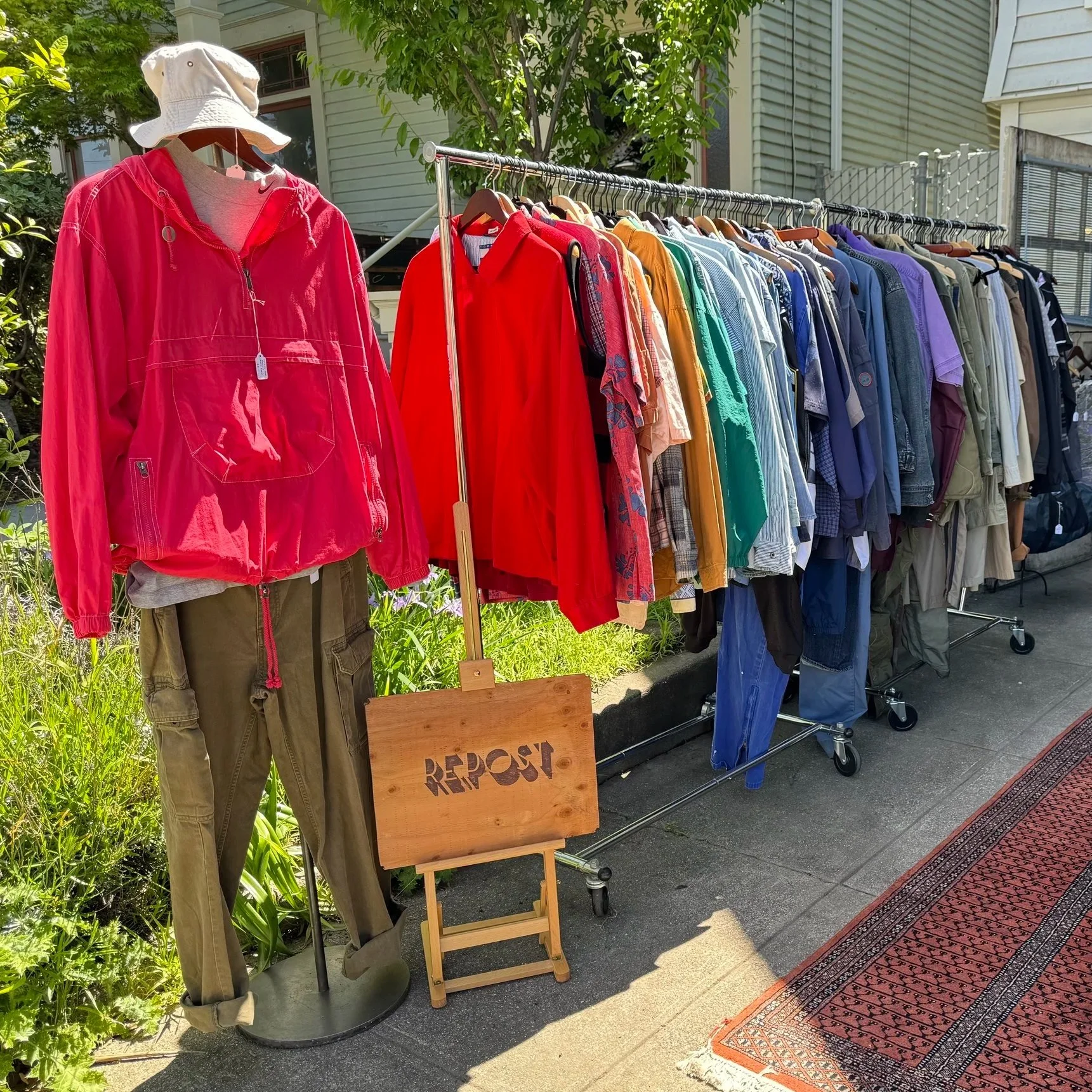 A clothing sale outside with a mannequin wearing khaki pants, a gray shirt, a bright pink jacket, and a white bucket hat. A clothing rack displays a variety of colorful shirts and jackets, with a wooden sign that says 'REPOST' in front of it. There i