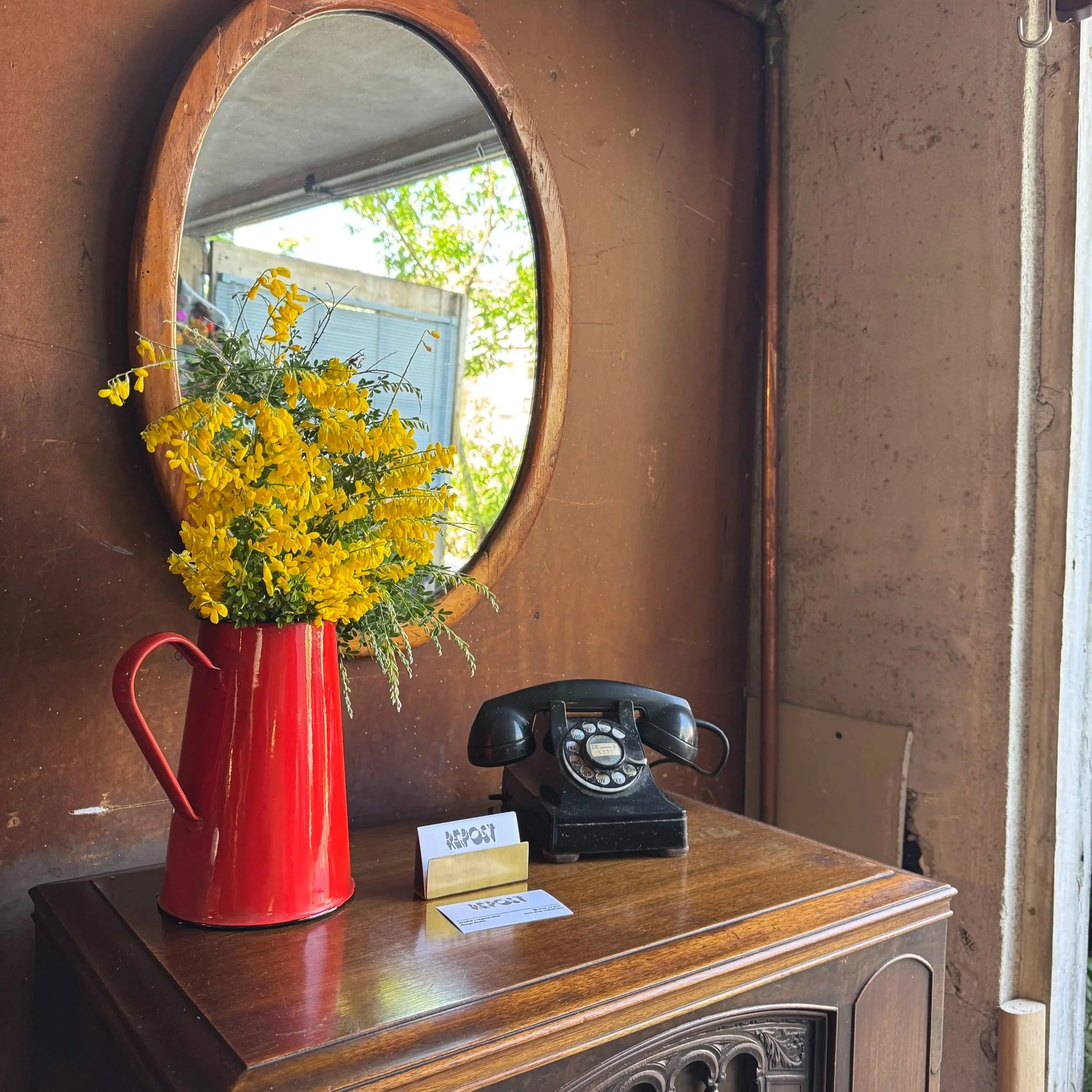 A decorative round mirror hung on a rustic brick wall above a wooden cabinet. There is a red watering can with yellow flowers sitting on the cabinet. To the right of the mirror, an old-fashioned rotary dial telephone and a folded receipt or paper are