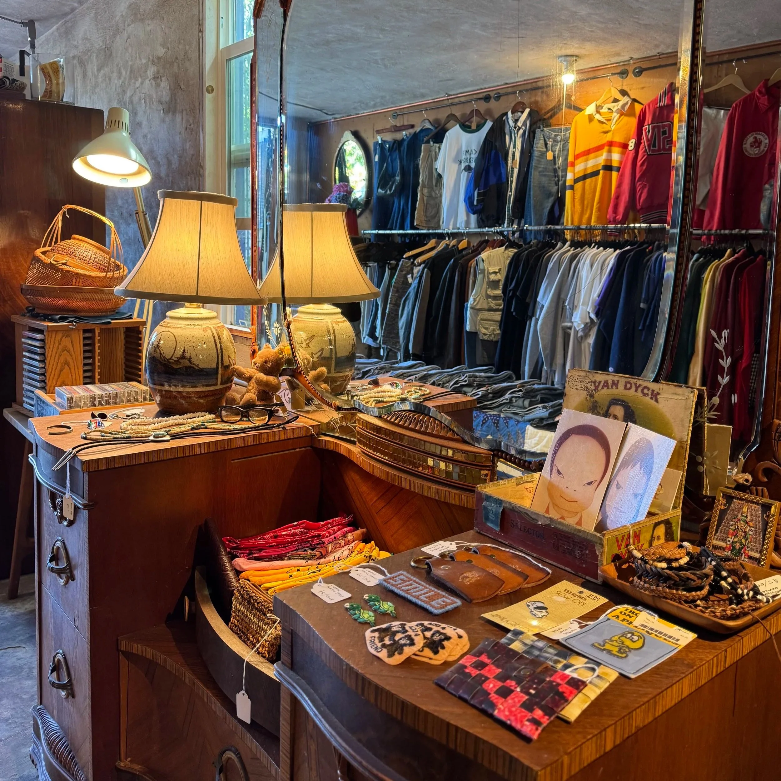 Display of vintage and secondhand items in a thrift store, including clothing on racks, pottery, jewelry, and small decorative objects on tables, with lamps illuminating the items.