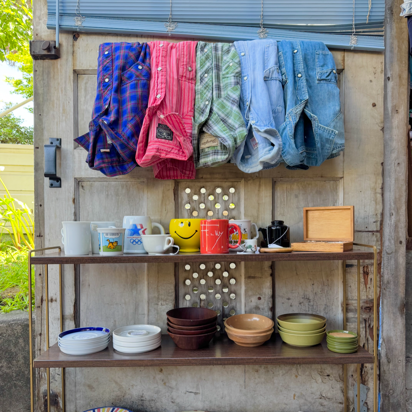 Clothes hang on a line above a shelf with mugs and bowls in a rustic outdoor setting.