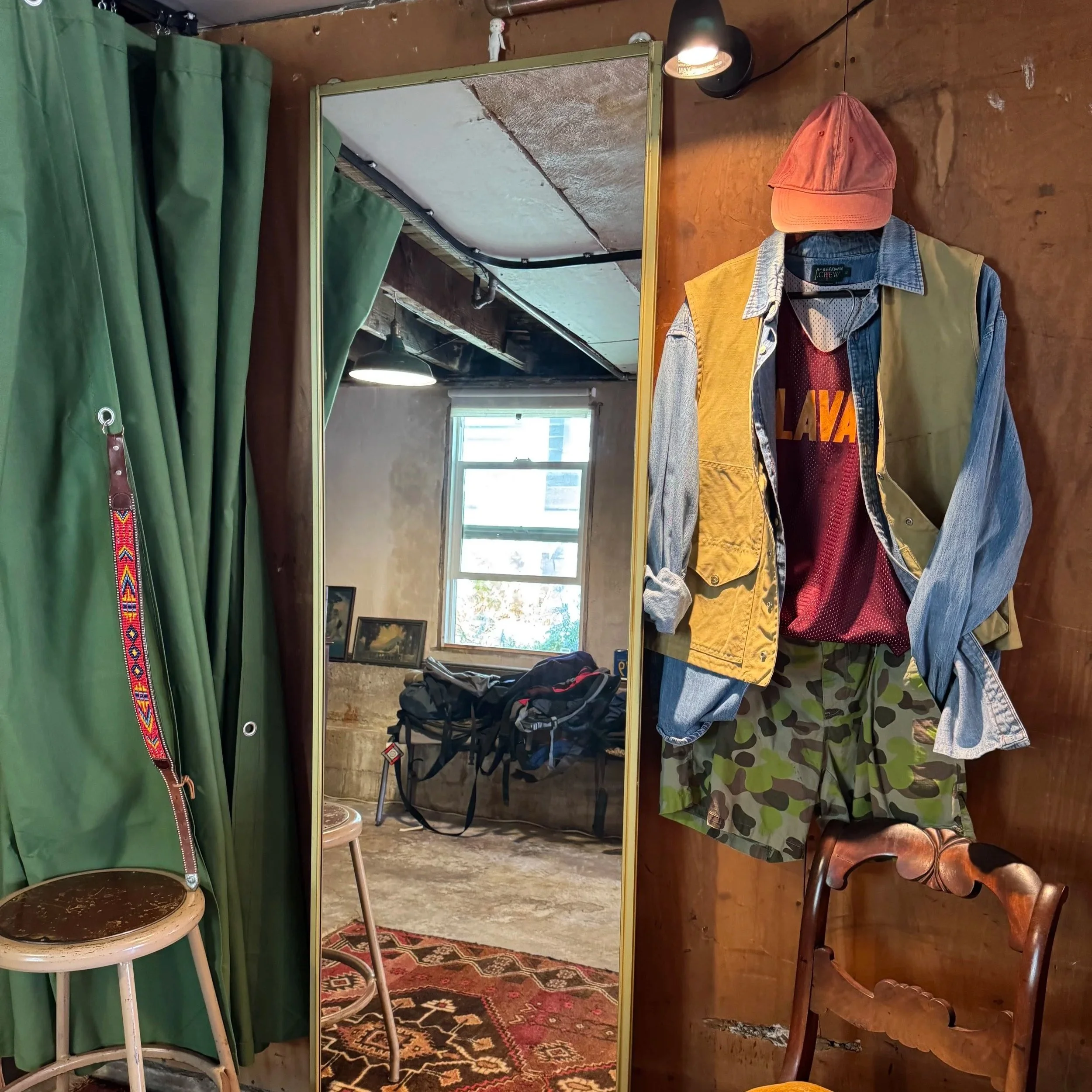 A large rectangular mirror reflecting a room with a window, wooden wall, and ceiling. In front of the mirror, there are casual clothes including a denim jacket, camouflage shorts, and a red shirt. To the right of the mirror, there is an orange cap, a