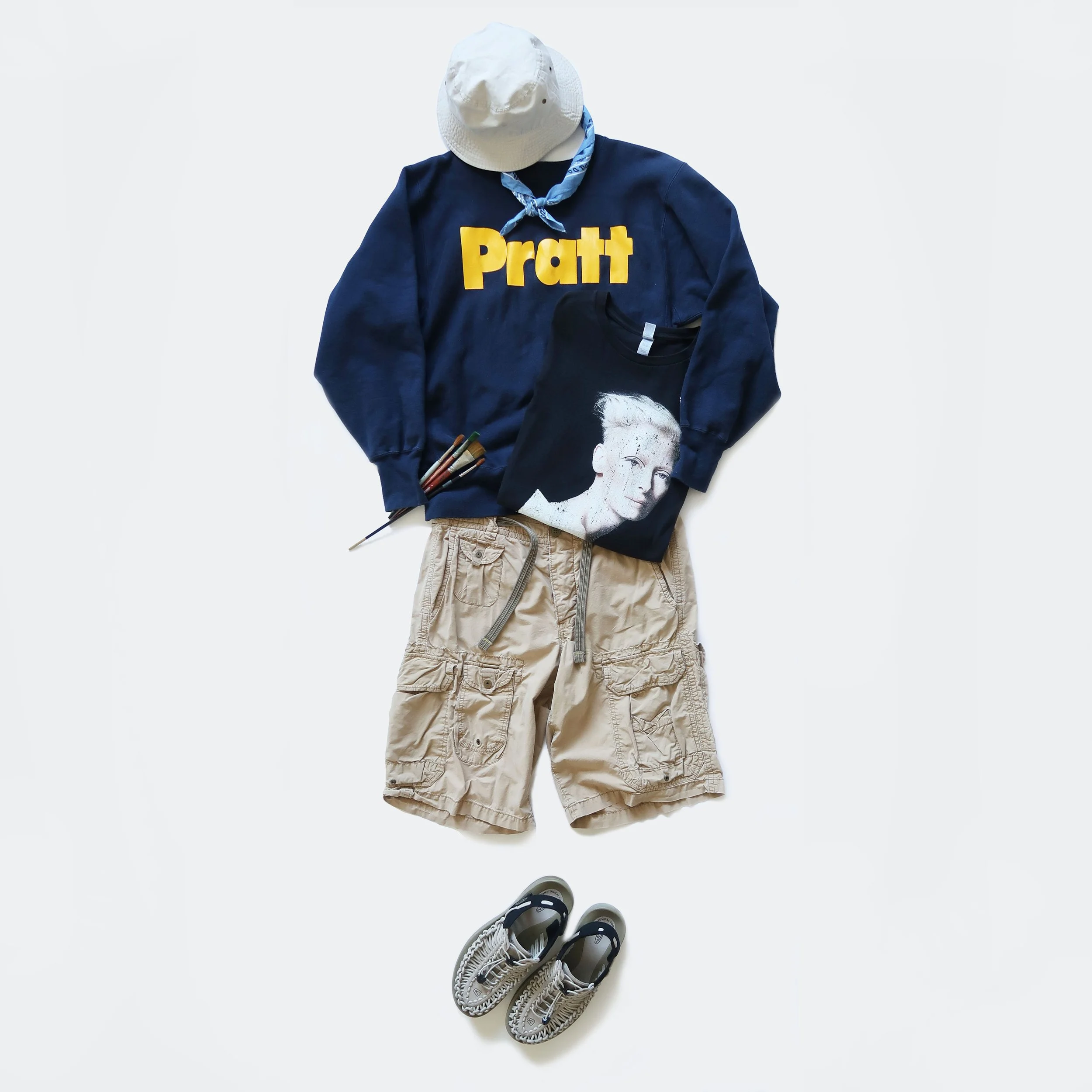 Casual outfit laid out, including a white bucket hat, blue sweatshirt with yellow text, beige cargo shorts, black and white sneakers, and a few markers.